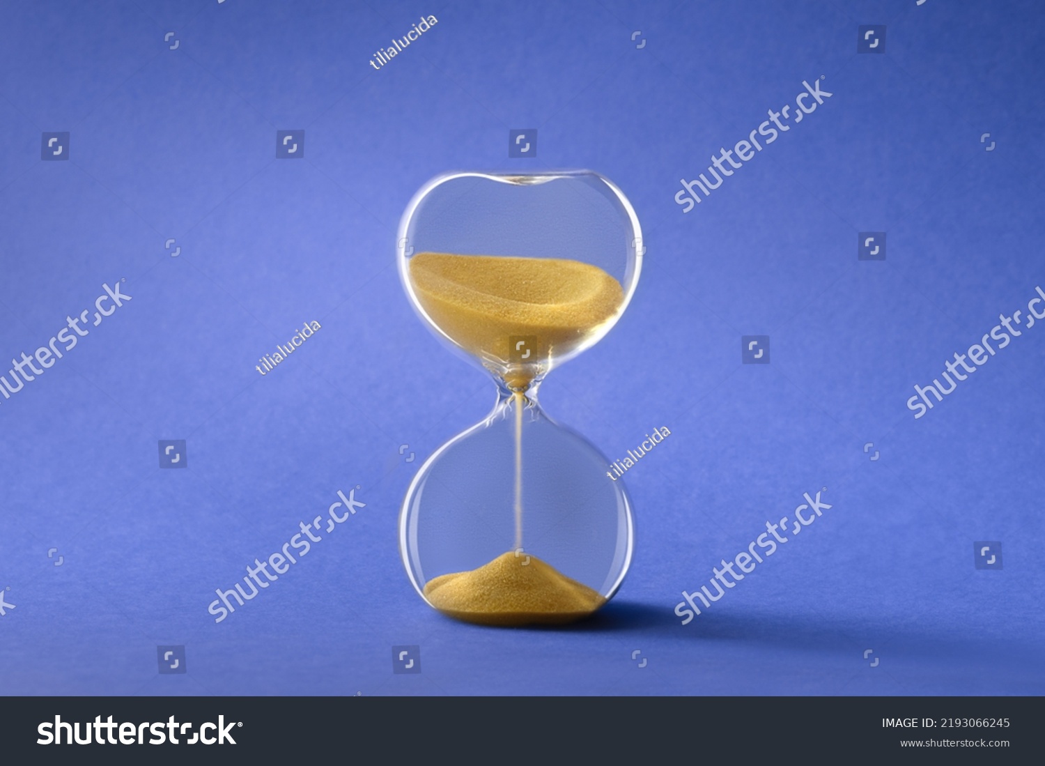 Hourglass, also known as sandglass or sand timer. Single glass sand clock with golden sand on blue paper background. Design element, #2193066245