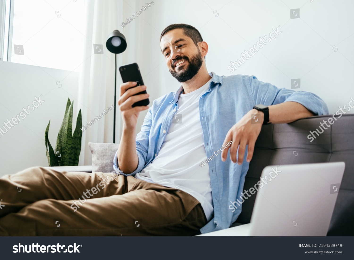 Smiling man sitting on couch at home using laptop and cell phone #2194389749