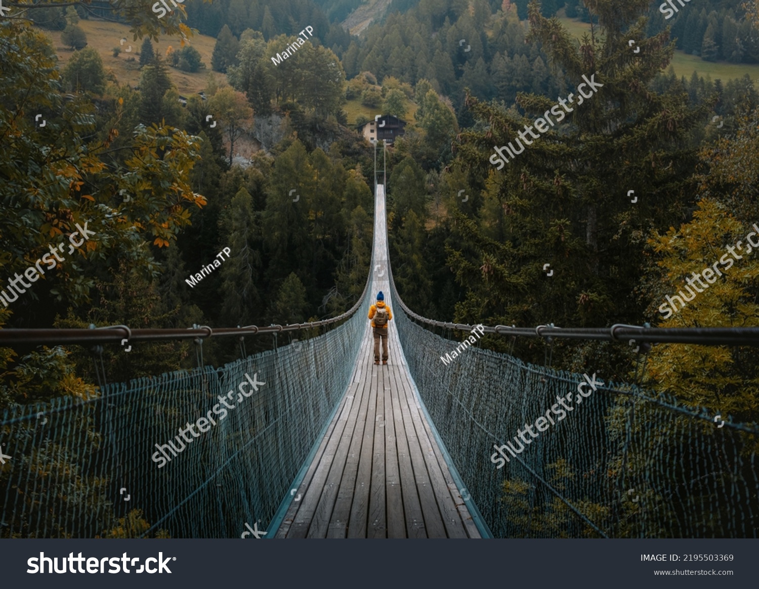 Young traveler man dressed in yellow jacket crosses hiking on an impressive wooden and metal bridge in the village of Goms in the Swiss alps #2195503369