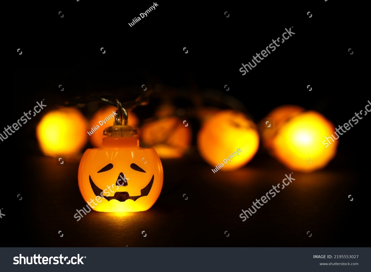 Garland lights of glowing halloween orange pumpkins with copy space above and below on black background. Blurred bokeh lights, one pumpkin face focused, close up. Festive holidays Halloween concept. #2195553027