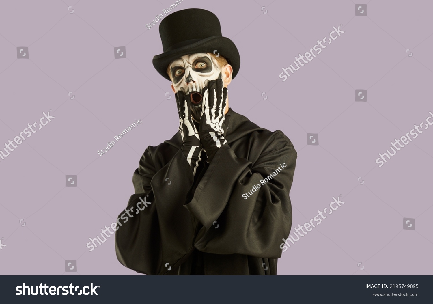 Portrait of shocked man in Halloween make-up and costume grabbing his face in fright. Man in black hat, suit and skull make-up opens his eyes and mouth wide in fear on light lilac background. #2195749895