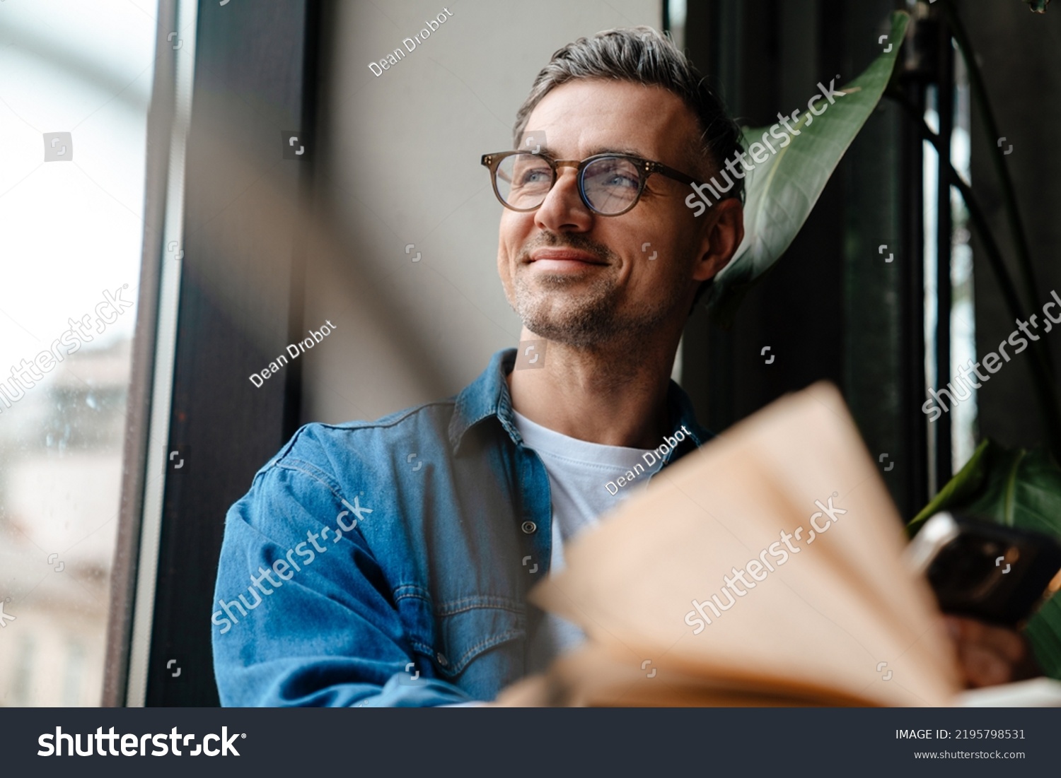 Portrait of adult handsome smiling man in glasses looking out the window while sitting with book in cafe #2195798531