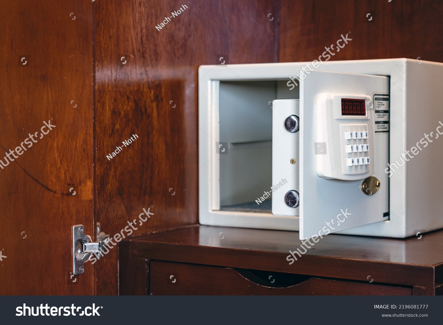 Security open metal safe with empty space inside in a wooden shelf. White safe box open door. Safe box with electronic lock in the hotel or home. Selective Focus on locking mechanism of small safes. #2196081777