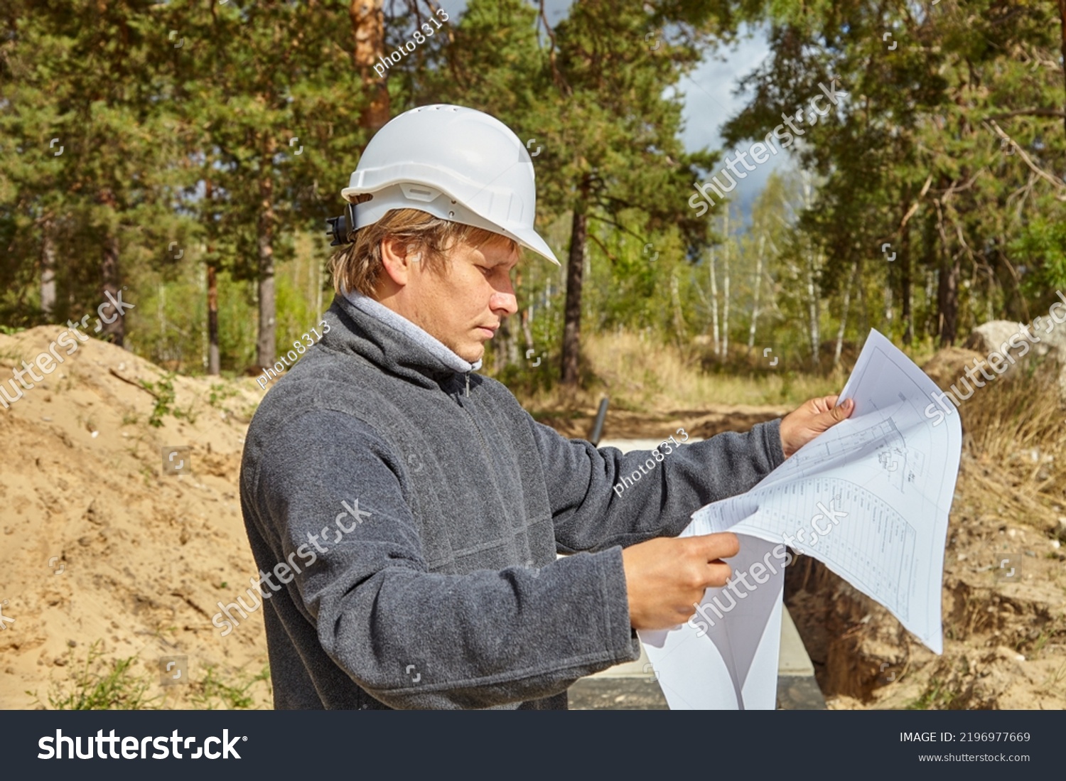 Engineer with a drawing on the construction site. A man in a white helmet. The engineer looks at the drawing. #2196977669