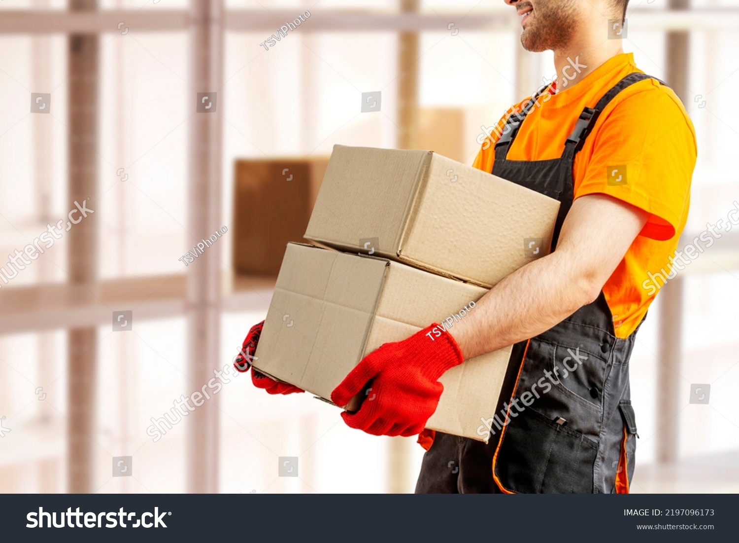 Young man holding cardboard package working in warehouse among racks and shelves. Delivery man with box. Staff laborer, orange uniform cap, t-shirt, coveralls service moving delivering orders goods.  #2197096173