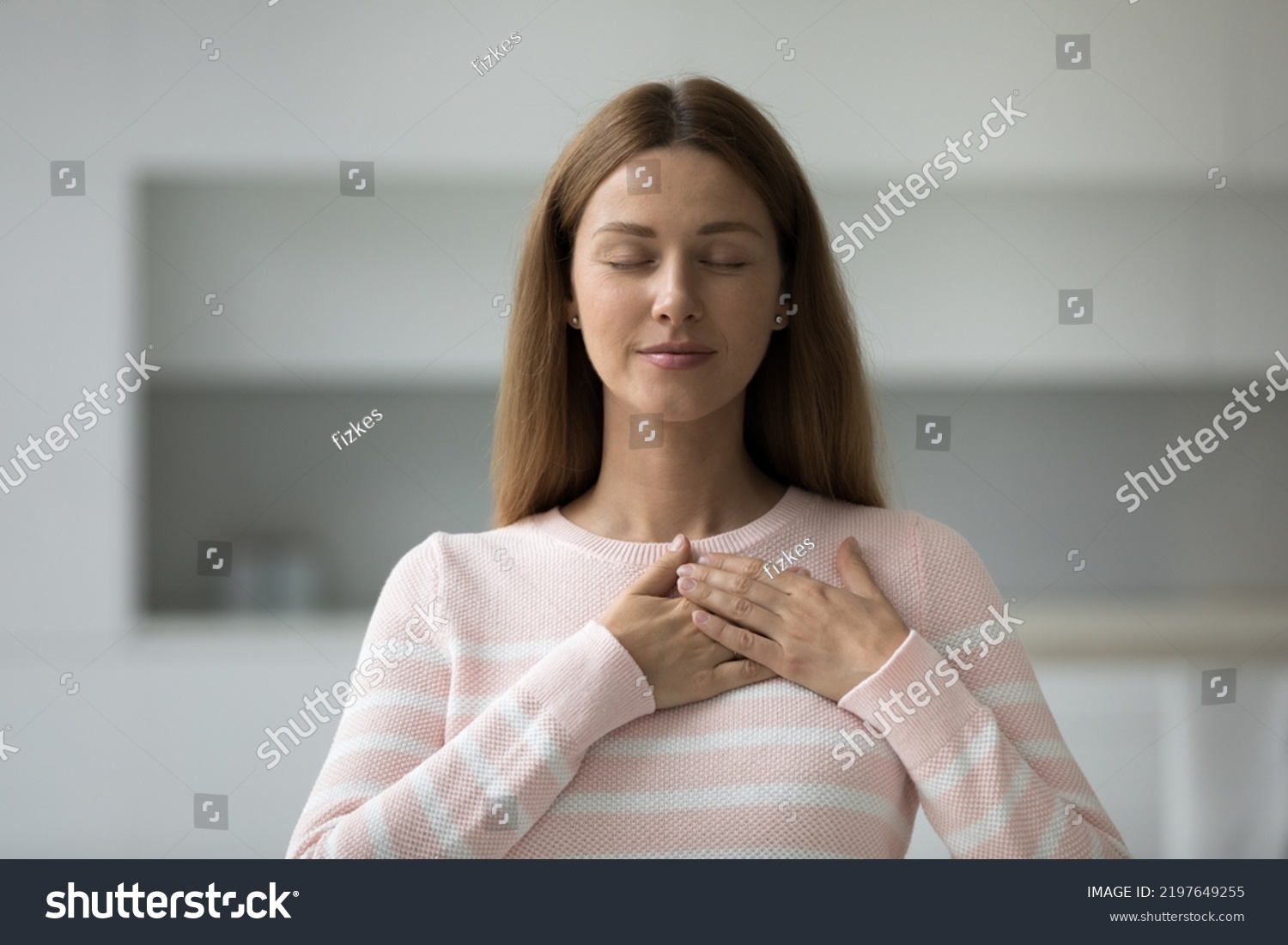 Peaceful beautiful Caucasian woman touching chest with both stacked hands. Grateful female model expressing gratitude, love, recognition. Pray, religion, faith #2197649255