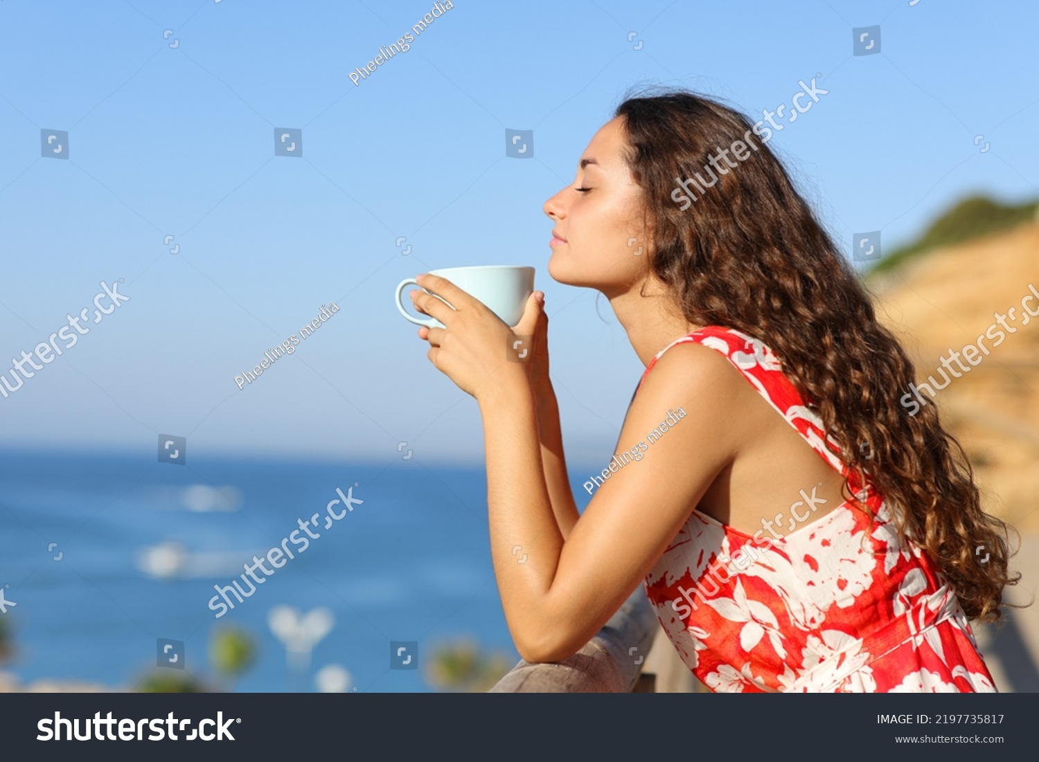 Side view portrait of a woman in red dress relaxing smelling coffee on the beach #2197735817