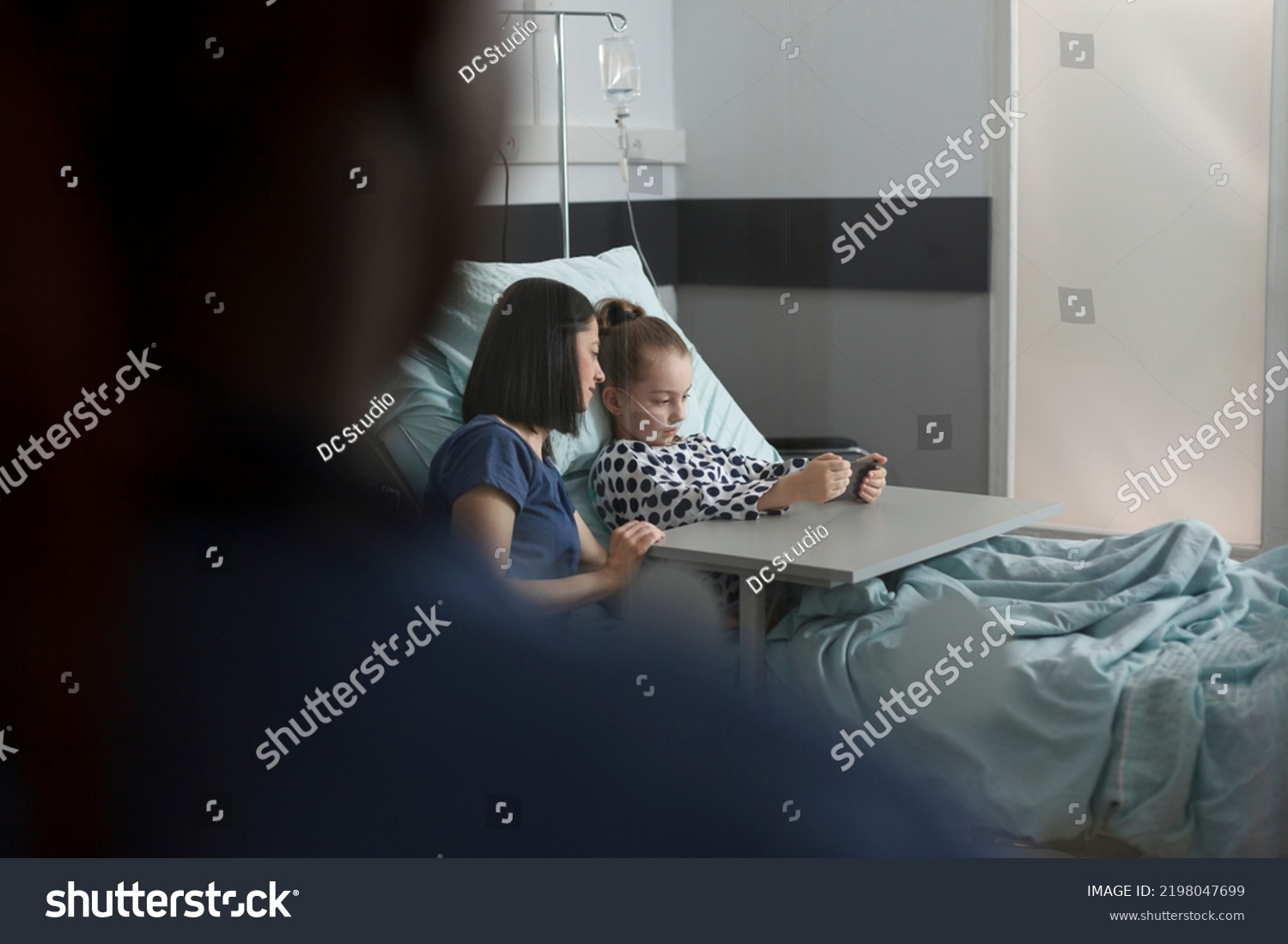 Caring parent sitting beside hospitalized sick daughter watching cartoons on smartphone. Ill little girl playing games on mobile phone while resting in hospital pediatrics ward patient bed. #2198047699