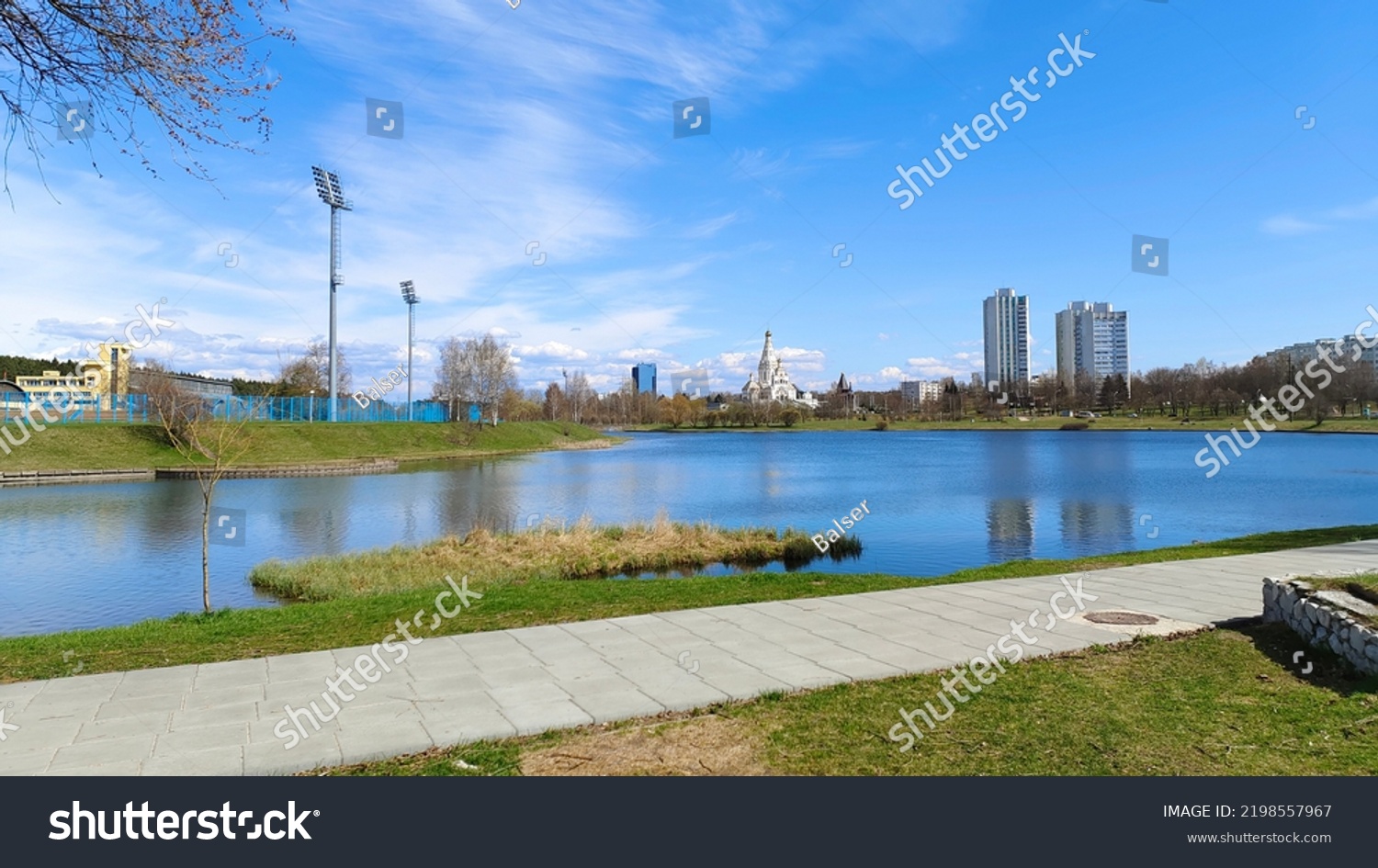 There are trees and grass on the shore of the lake. Along the shore is a paved sidewalk. On the opposite shore is a stadium with lighting poles, an Orthodox church and apartment buildings. Sunny #2198557967