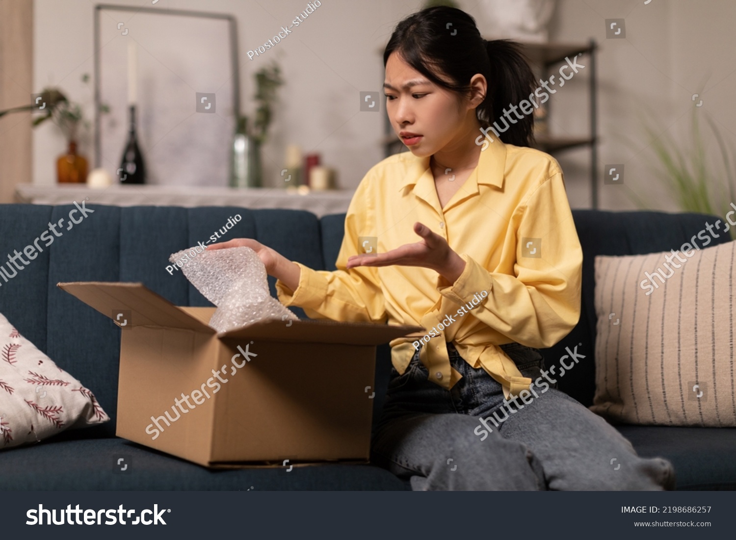 Discontented Asian Lady Buyer Unpacking Cardboard Box With Bad Product Sitting On Sofa At Home. Negative Feedback, Delivery Service And Shopping Problem Concept #2198686257