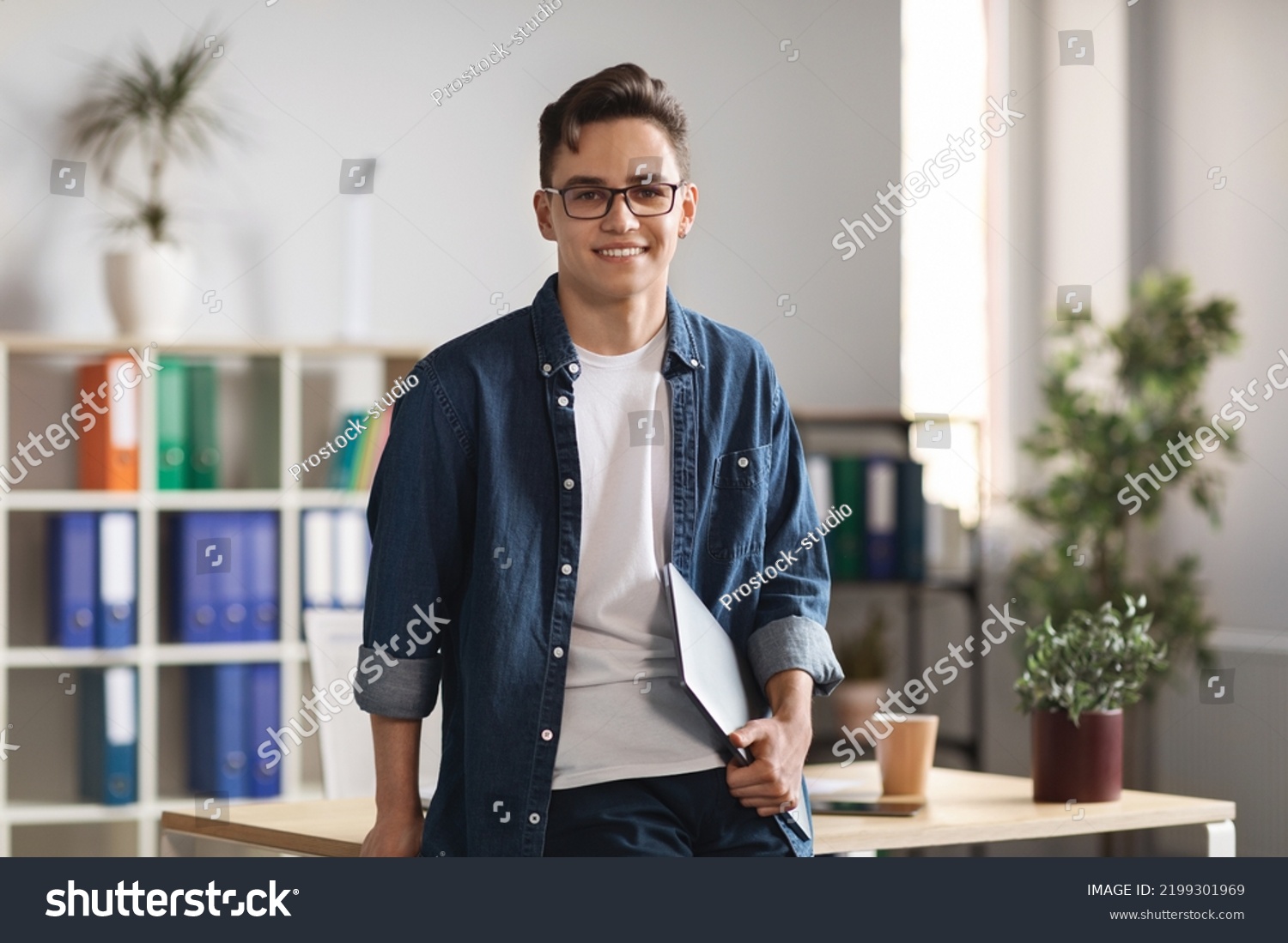 Portrait Of Handsome Young Male Entrepreneur Holding Laptop And Posing In Office Interior, Confident Millennial Man In Casual Clothes Standing Near Desk And Smiling At Camera, Copy Space #2199301969