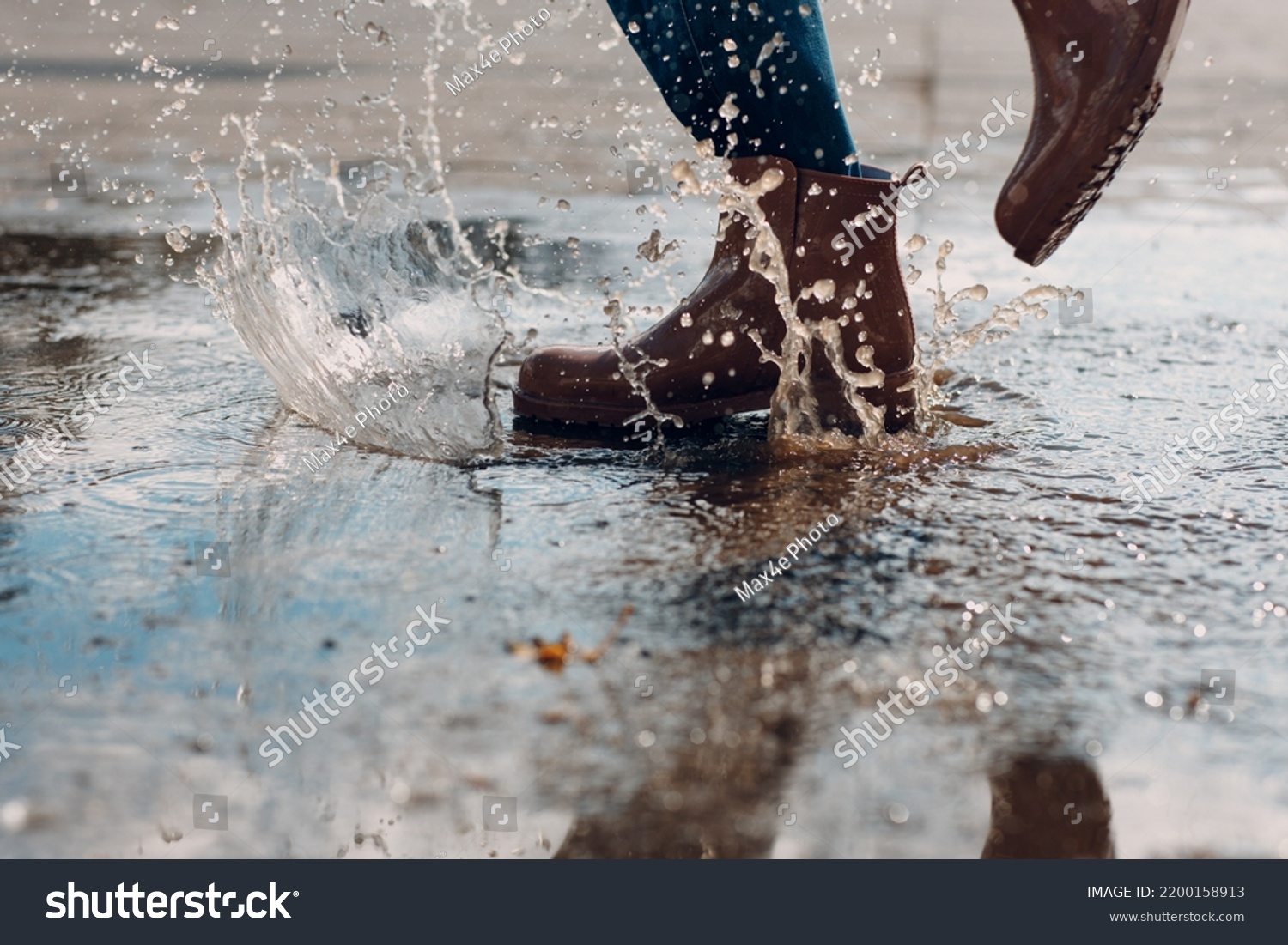 Woman wearing rain rubber boots walking running and jumping into puddle with water splash and drops in autumn rain. #2200158913