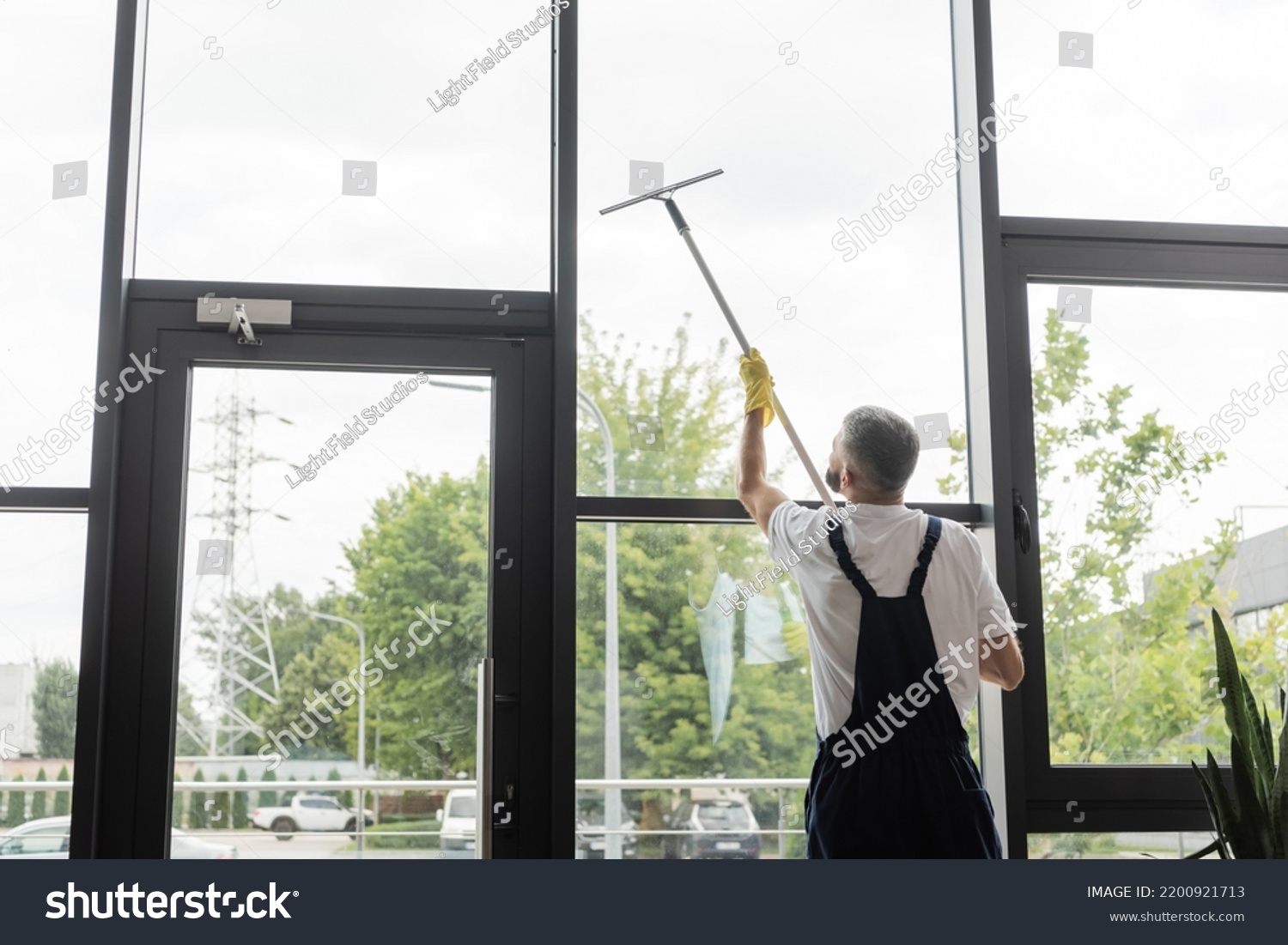 back view of man in overalls washing large office windows with window squeegee #2200921713