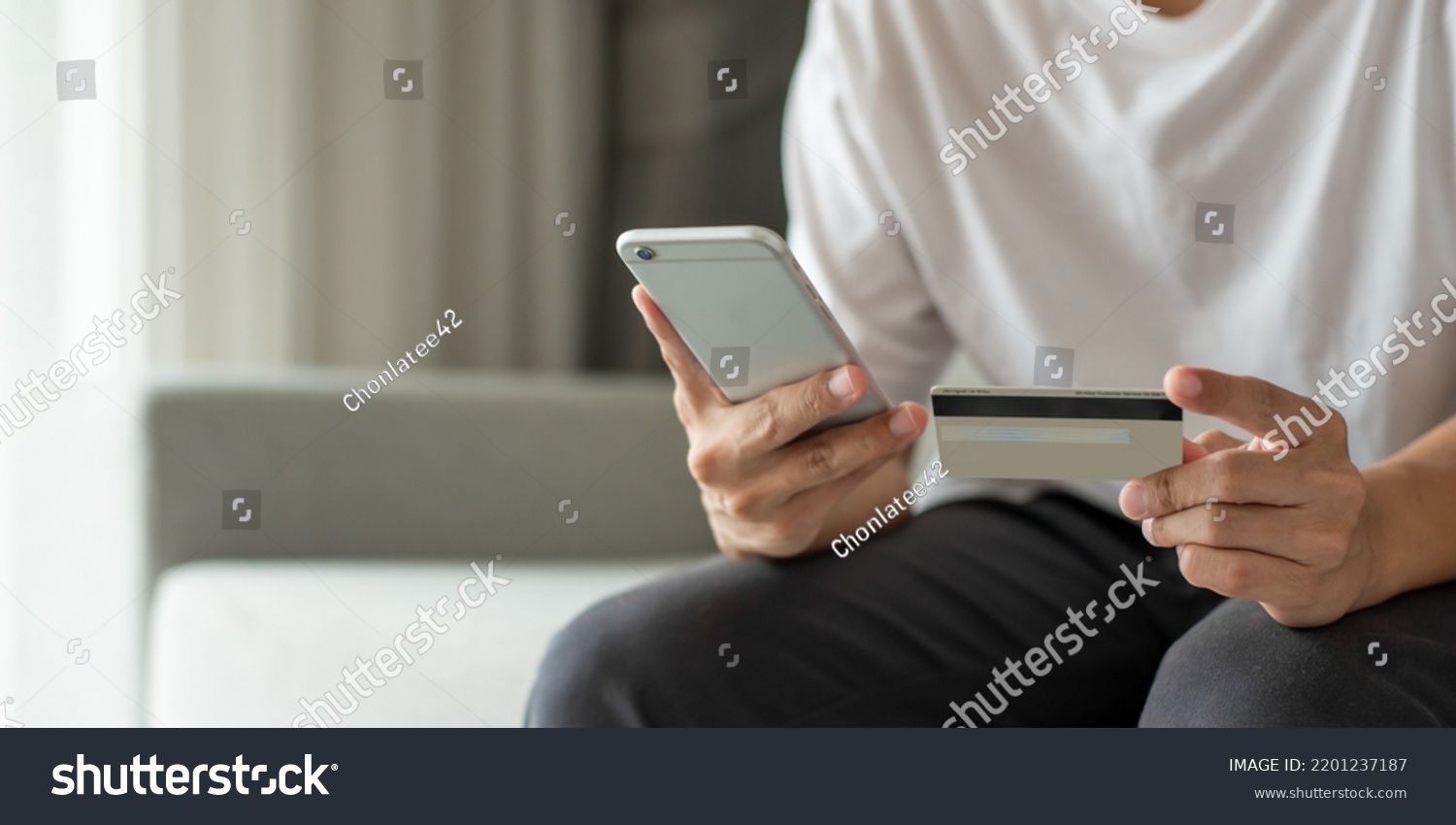 Young man in a white shirt sits and using smartphone and hand holding credit card for pay online shopping on the couch at home. She smiled happily not having to leave the house to go shopping. #2201237187
