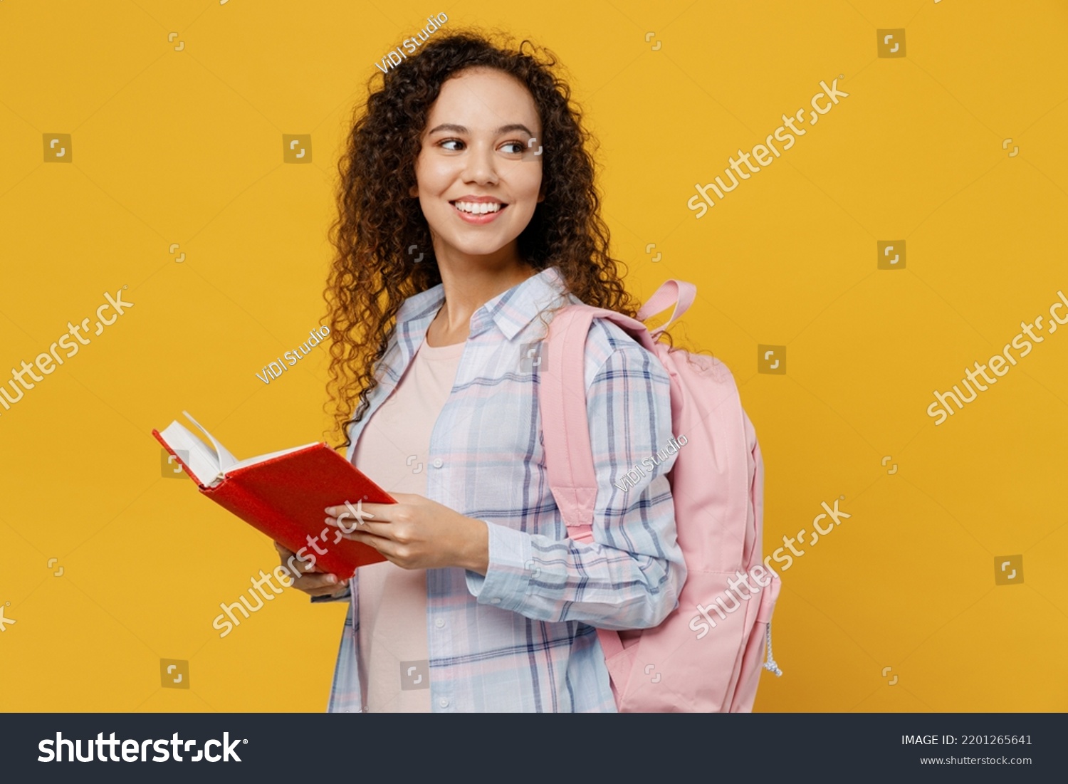 Side view young smiling happy black teen girl student she wear casual clothes backpack bag read book look aside on area isolated on plain yellow color background High school university college concept #2201265641