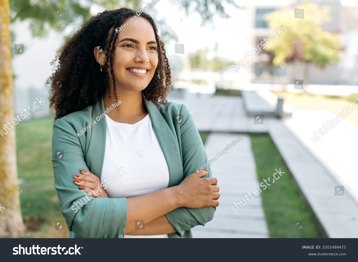 Portrait of lovely positive confident successful brazilian or latino woman with curly hair, business lady, in stylish elegant clothes, stands outdoors with arms crossed, looking ahead, smiles friendly #2201484475