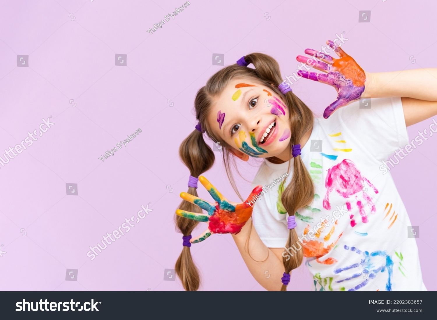 A beautiful little girl stained in multicolored paints on a pink isolated background has fun smiling. #2202383657