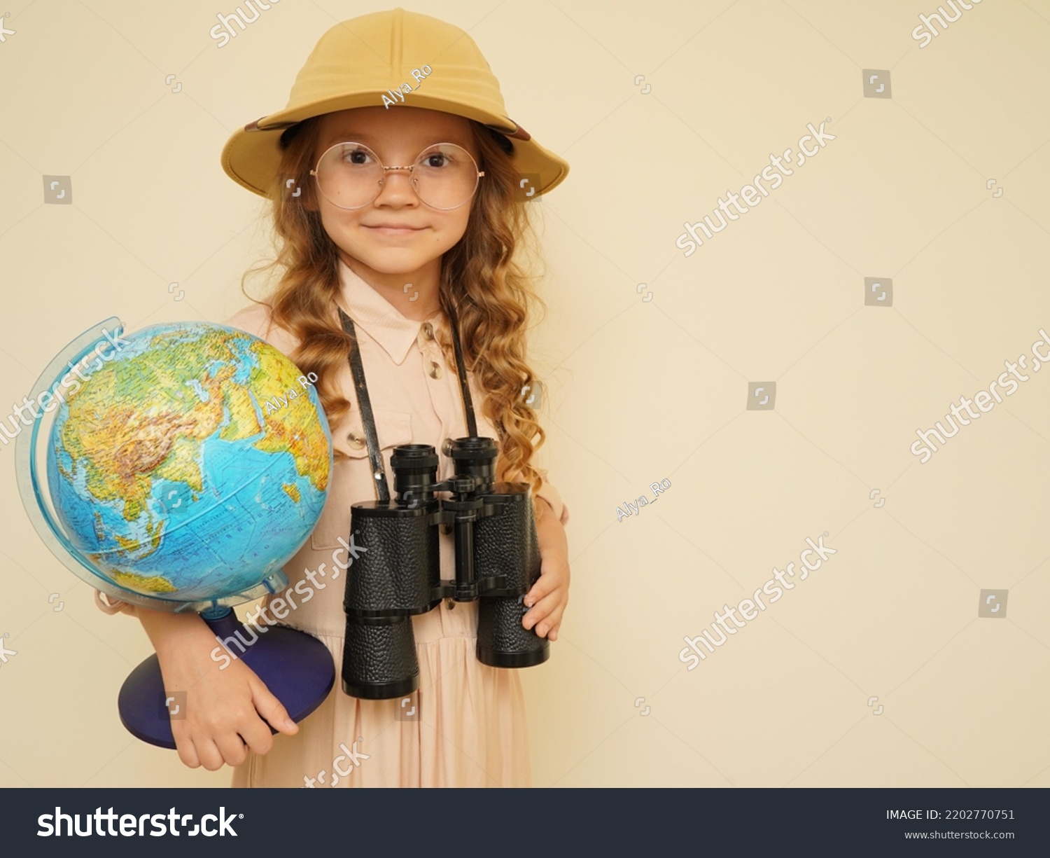 Happy child plays the discoverer. Unique child. Travel, adventure, vacation and discovery concept. A kid in a traveler's hat with binoculars and a globe in his hands on the beige background.  #2202770751
