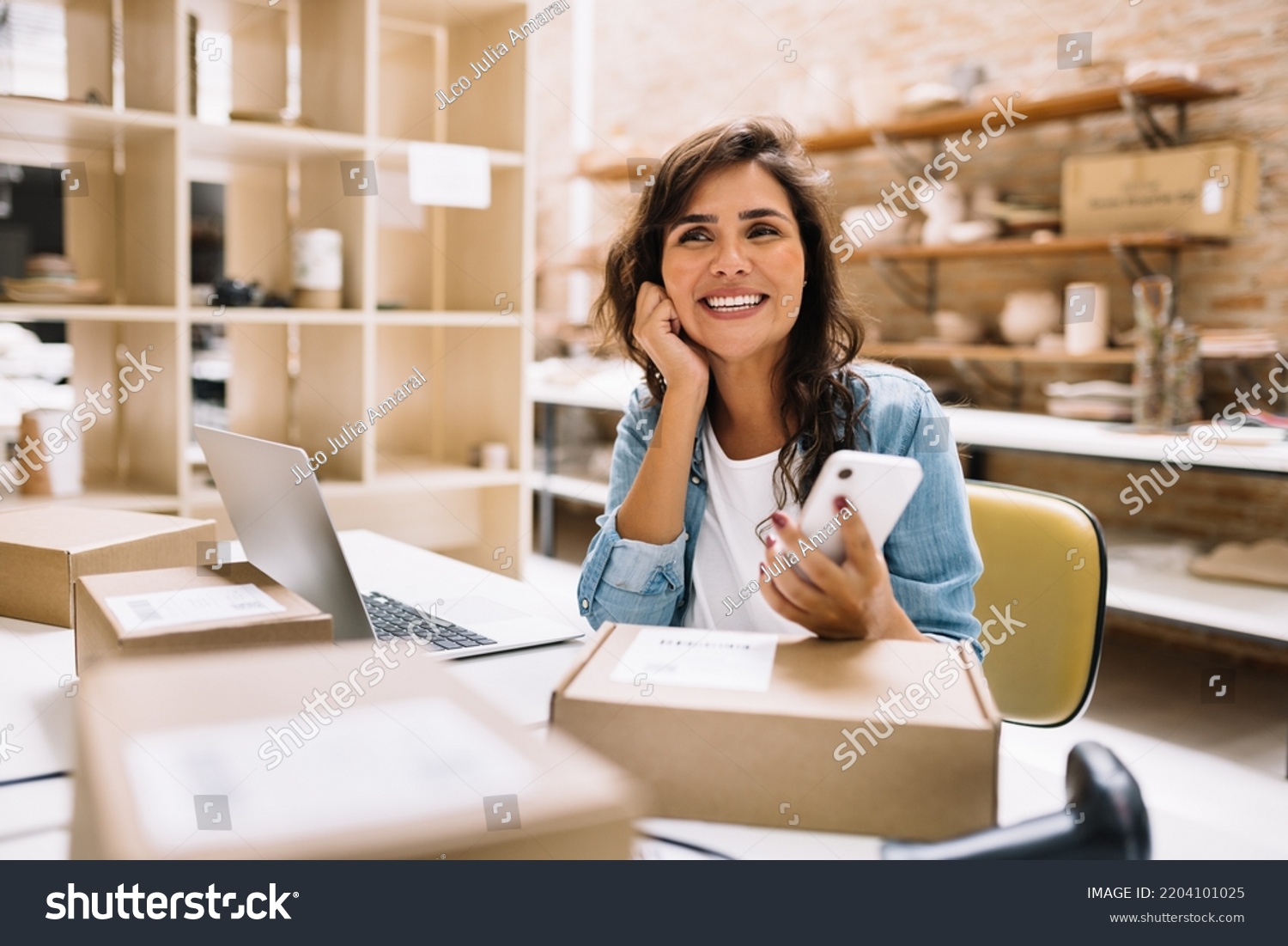 Cheerful online store owner looking away thoughtfully while holding a smartphone. Female entrepreneur preparing orders for shipping in a warehouse. Businesswoman running an e-commerce small business. #2204101025