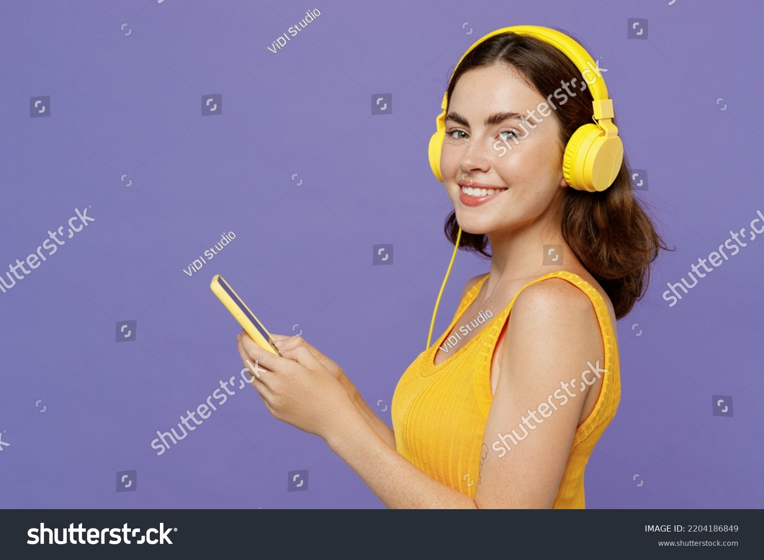 Side view young woman 20s wear yellow tank shirt headphones listen to music hold in hand use mobile cell phone look camera isolated on plain pastel light purple background. People lifestyle concept #2204186849