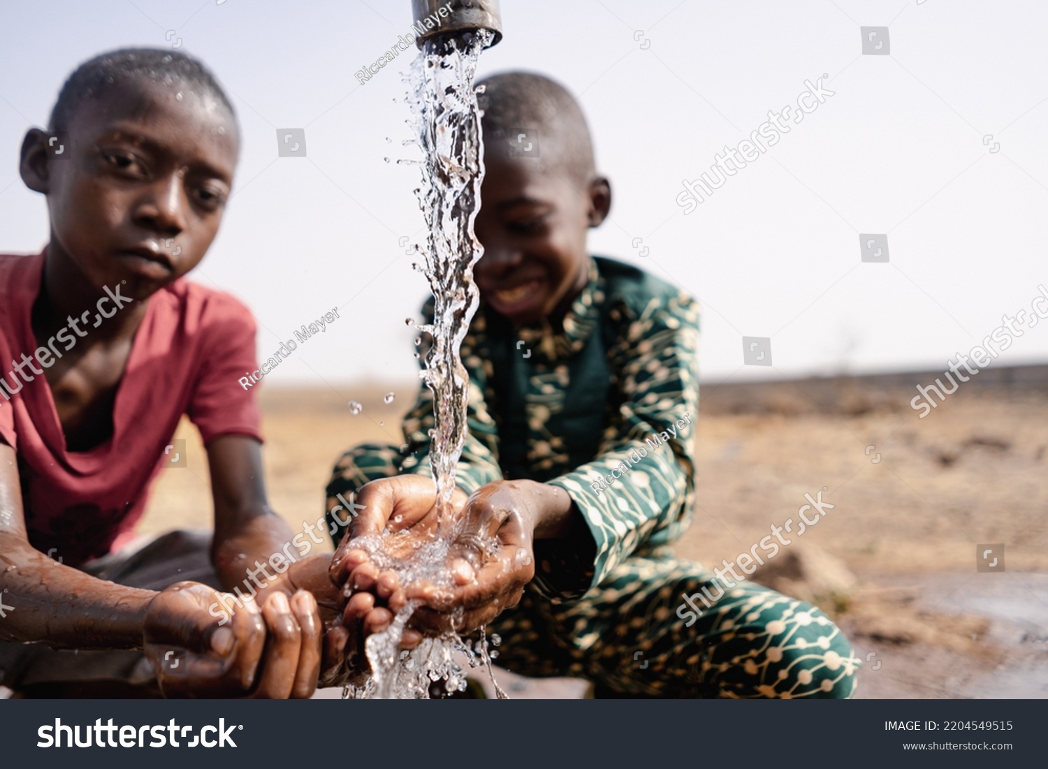 Two African toddlers playing with the water that flows from a rural faucet on the outskirts of their village; concept of water scarcity #2204549515