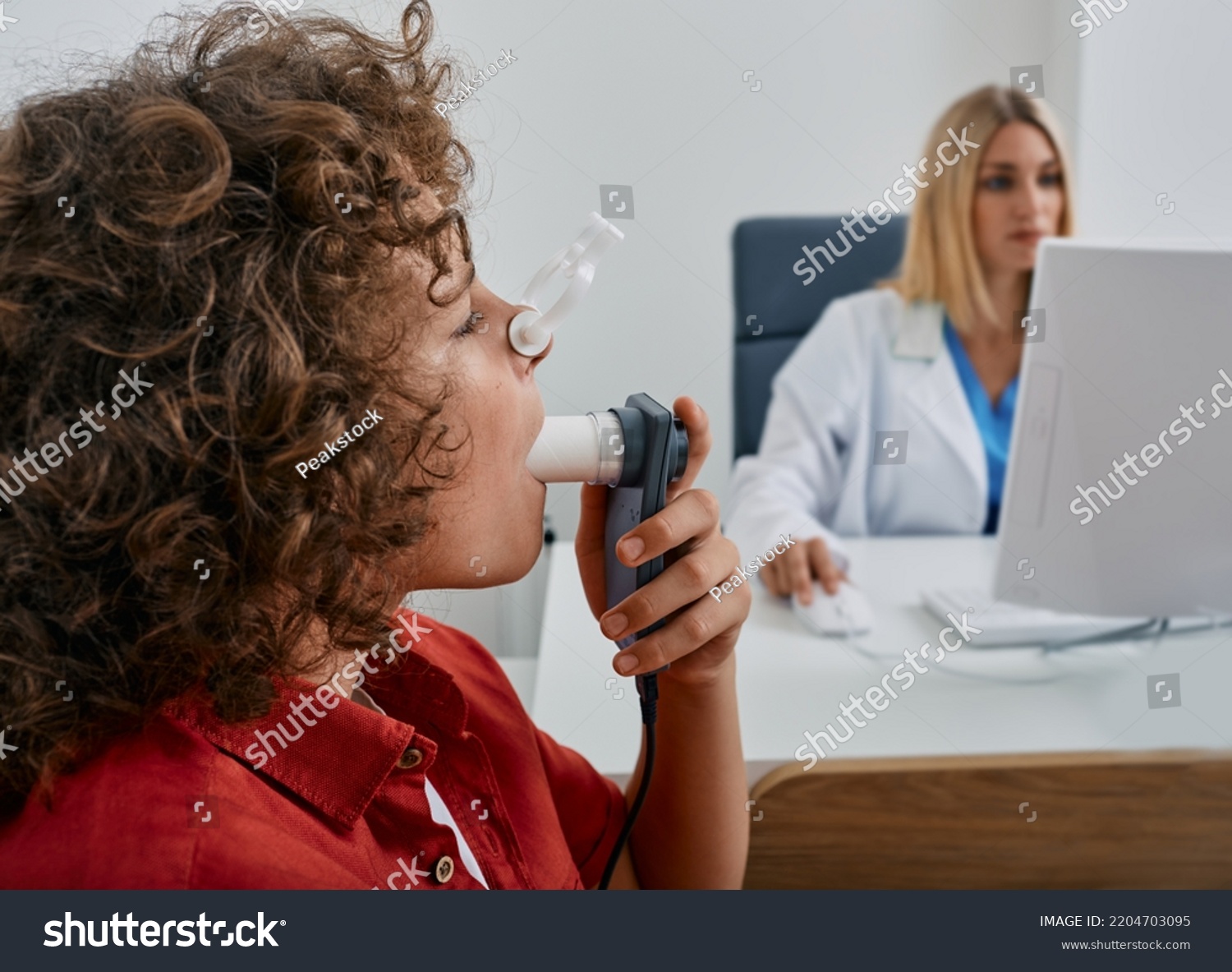 Male child during breathing spirometry and pulmonary function test using medical spirometer with doctor looking at test results on monitor #2204703095