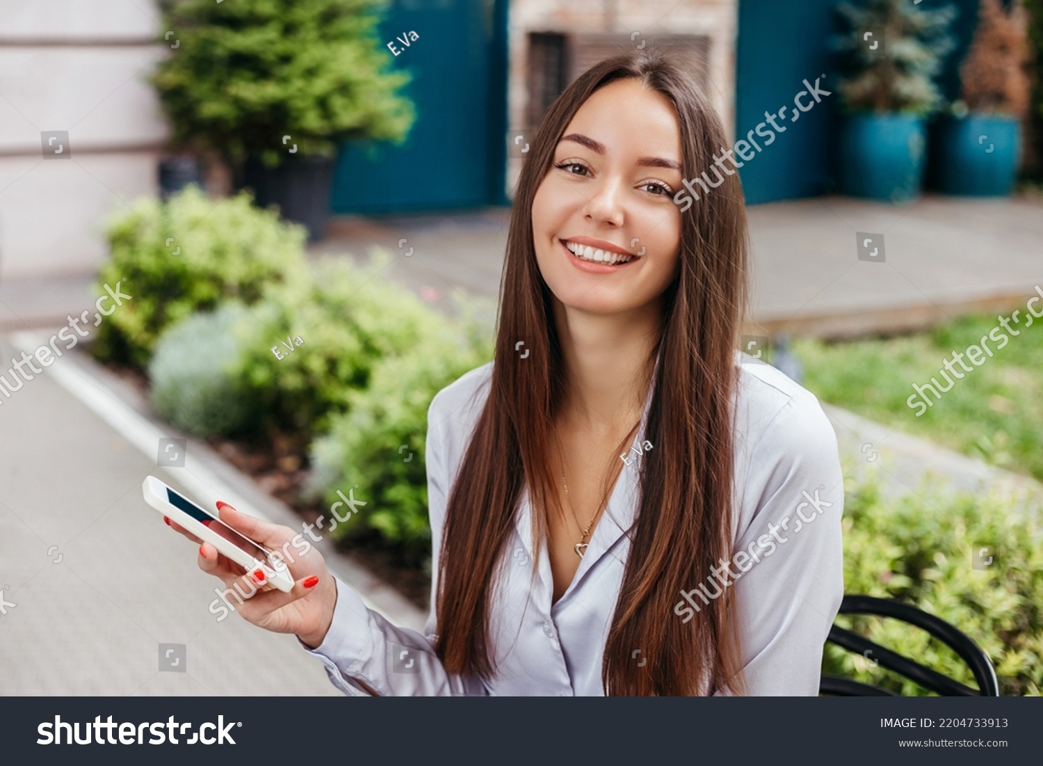 Portrait of a smiling girl with a mobile phone against the background of the facade of the cafe building. young brunette woman using a mobile phone and looking at the camera. Distant work #2204733913