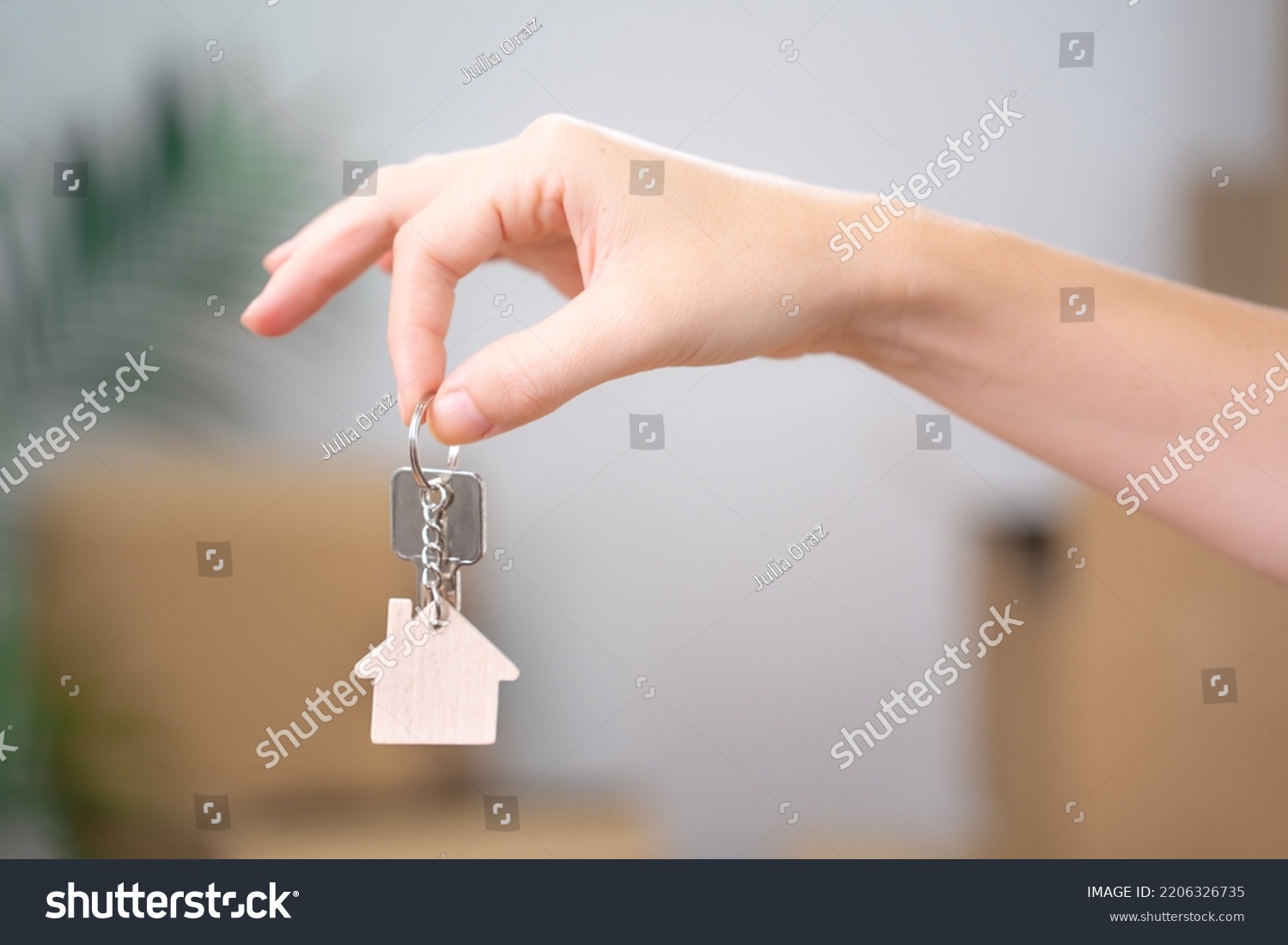 Female Hand Holding House Keys Inside Empty Room with Boxes Banner. Real estate agent handing over house keys in hand. Close-up view of keys from new home on cardboard box during relocation. #2206326735