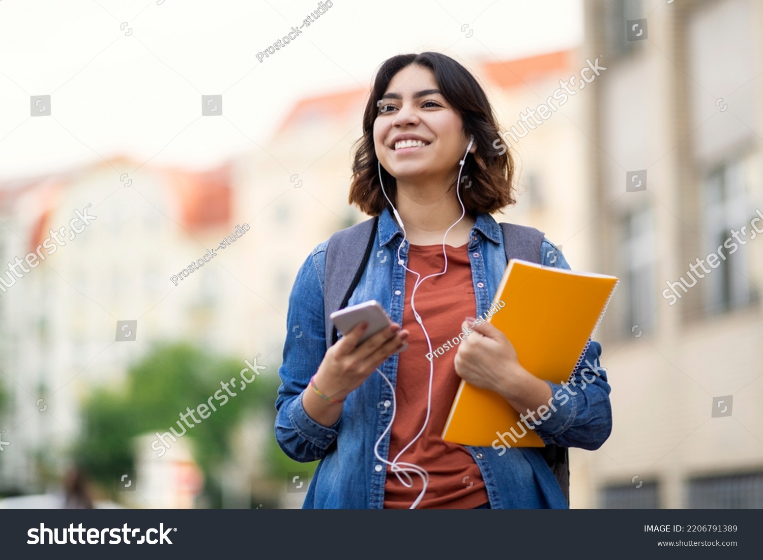 Cheerful young arab student female listening music with smartphone and earphones outdoors, smiling middle eastern woman carrying workbooks and backpack while walking on city street, copy space #2206791389