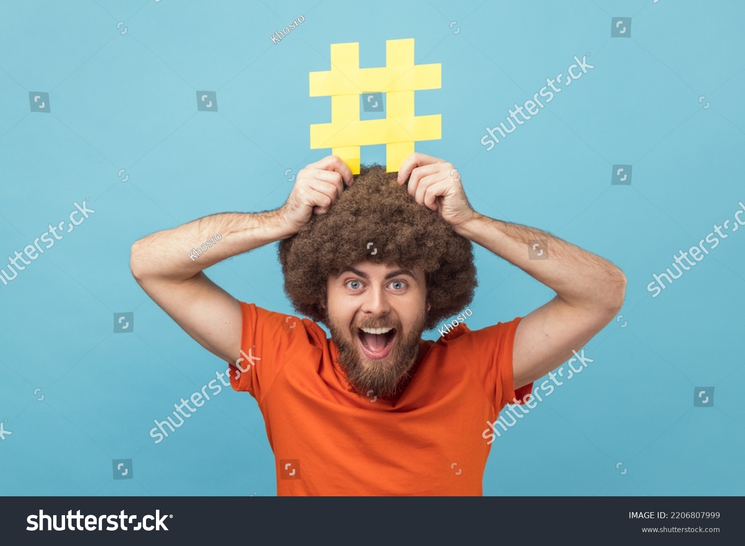 Portrait of man with Afro hairstyle holding yellow hashtag sign board on head, dreaming about target tagging posts on his website, smm manager. Indoor studio shot isolated on blue background. #2206807999