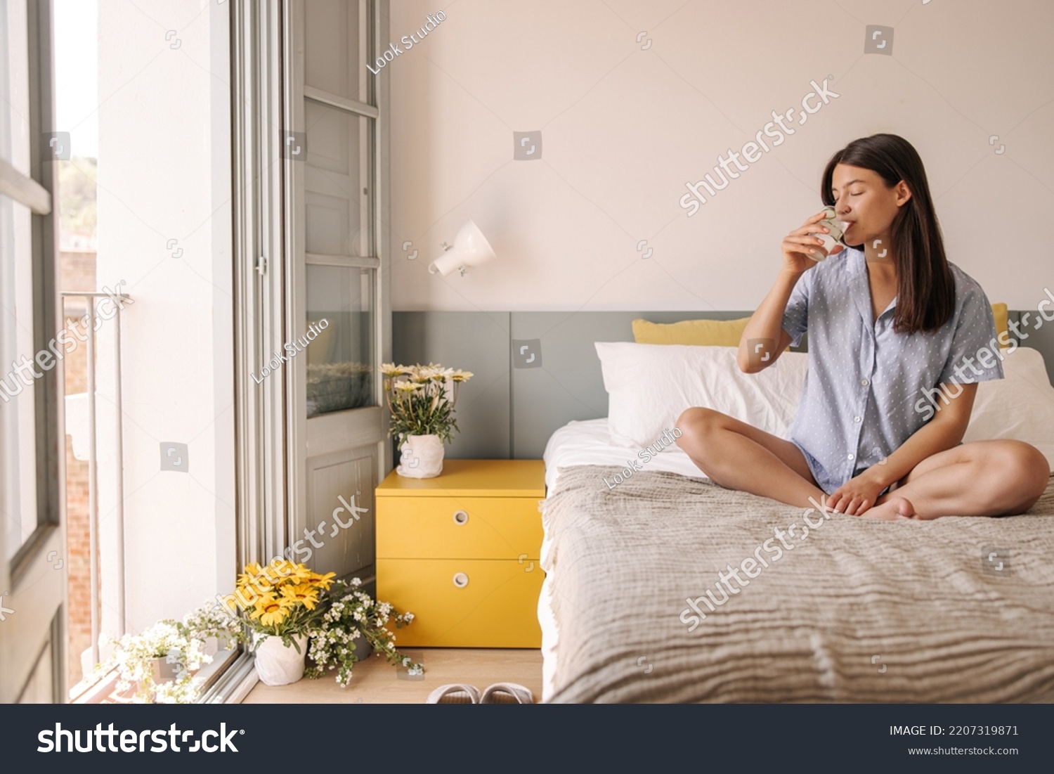 Nice young caucasian girl drinks water, crossing legs sitting on bed in room. Woman with brunette hair closes her eyes in pleasure. Good morning concept #2207319871