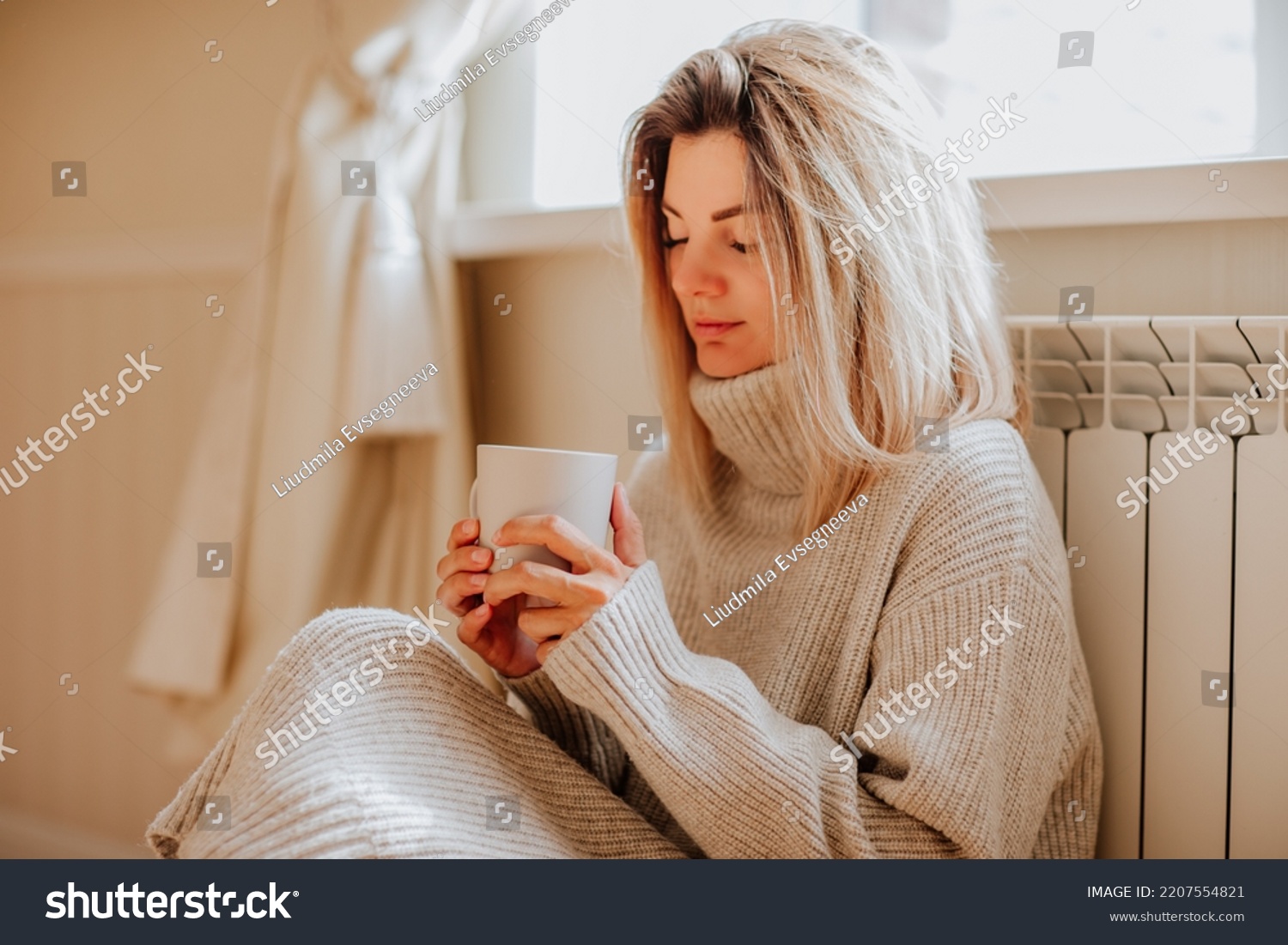 Young blond woman in long winter beige sweater is holding a cup of coffee and posing at home near the radiator. Winter season concept. Focus is at hands. Economy program #2207554821