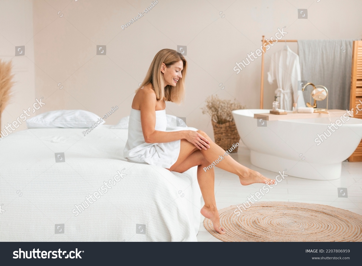 Beautiful middle aged woman in towel touching her leg with soft skin after depilation, sitting on bed at home, copy space. Mature lady after hair removal procedure, full length #2207806959