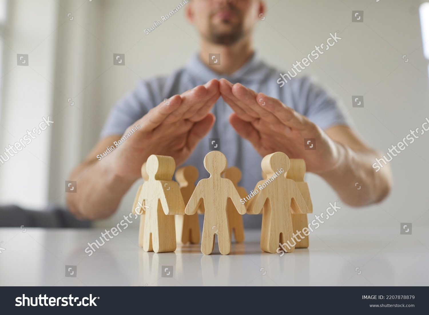 Young man holding hands above small wooden toy human figures placed on white desk as metaphor for human rights protection and safe community of people. Close up, closeup. Society, care, safety concept #2207878879