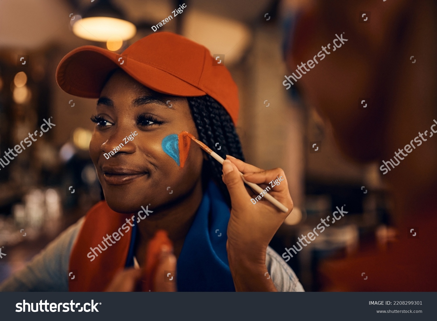 African American sports fan painting heart shape in team's colors on her face while getting ready for the match during soccer world cup.  #2208299301