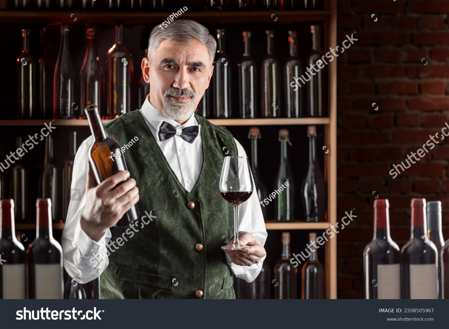 Sommelier with a glass of wine. Examination of wine products. Restaurant staff, expert wine steward among shelves of wine bottles. Stylish middle-aged man with a grey beard. #2208505967
