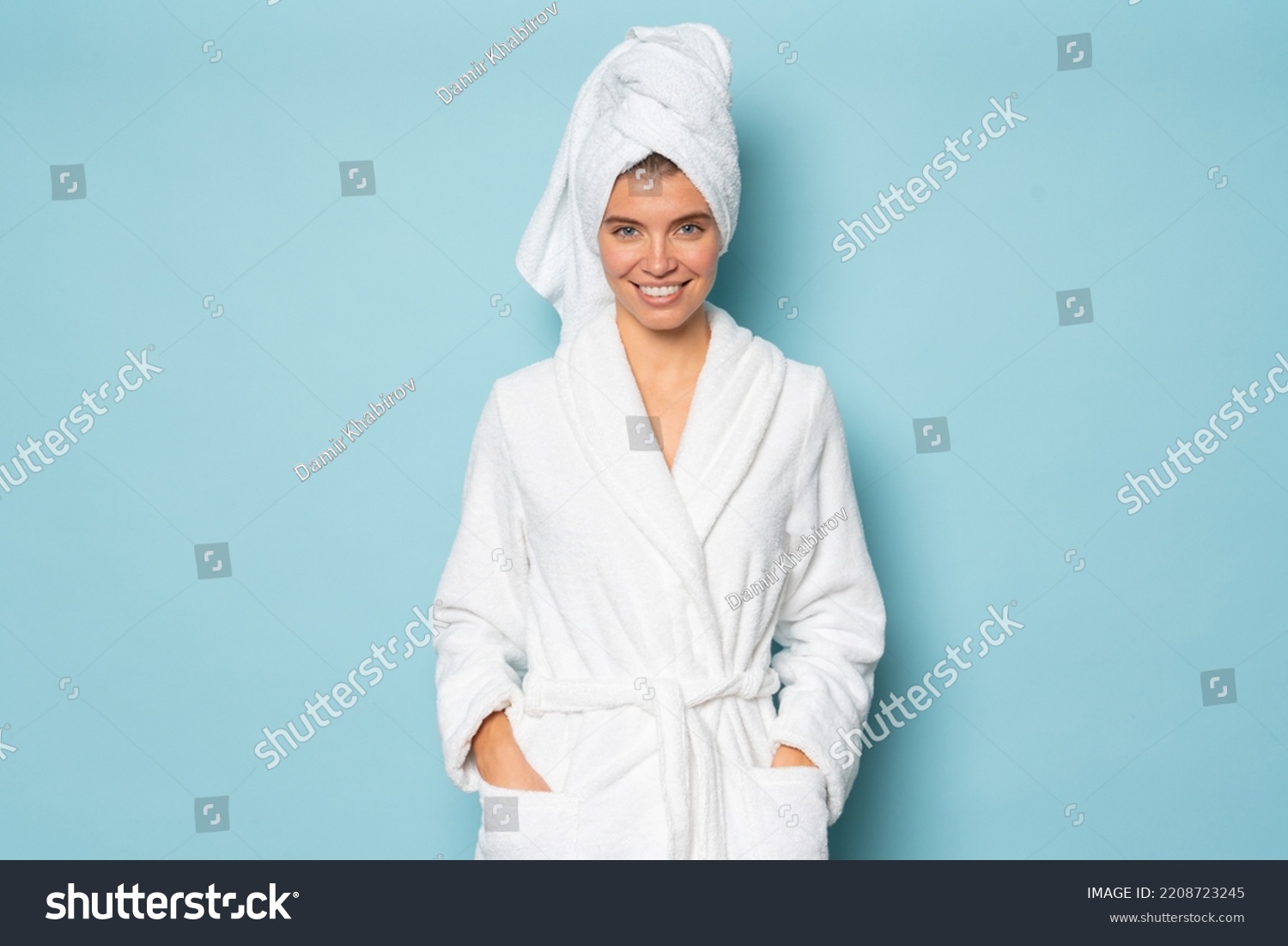 Smiling woman with clean body wearing white bathrobe and towel on head standing on blue studio background with hands in pocket at spa day on vacation or weekend. Hygiene, purity concept #2208723245