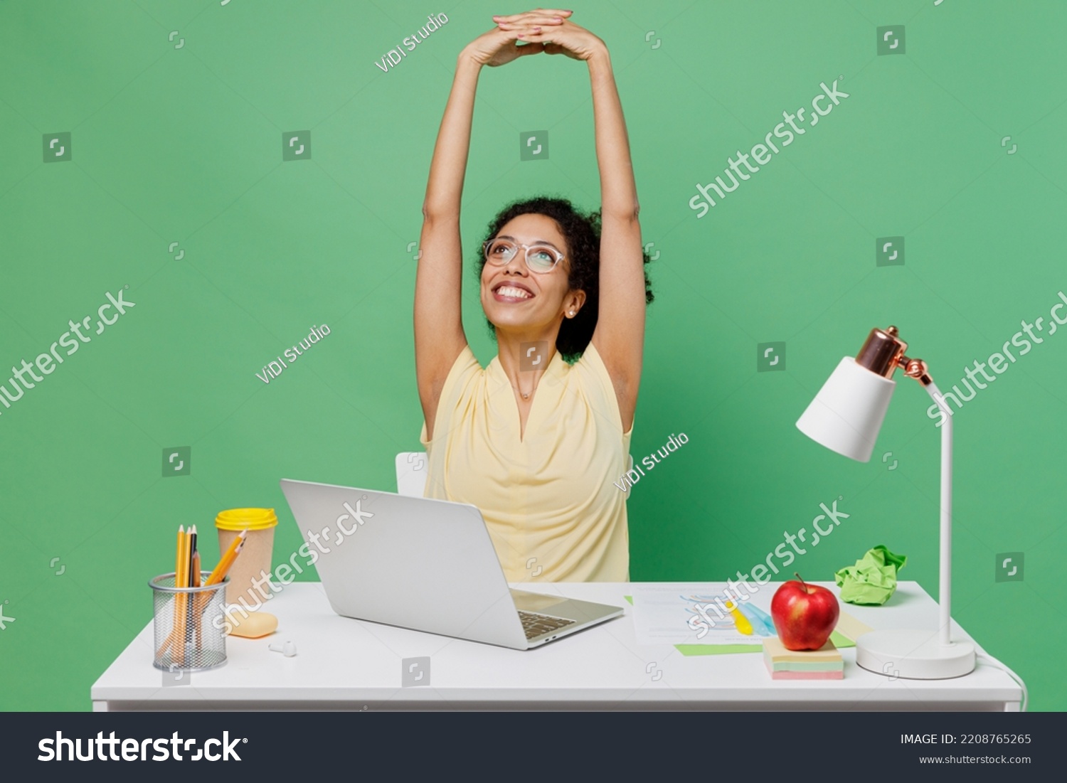 Young employee business woman of African American ethnicity wear shirt sit work at white office desk with pc laptop stretch hands rest isolated on plain green background. Achievement career concept. #2208765265