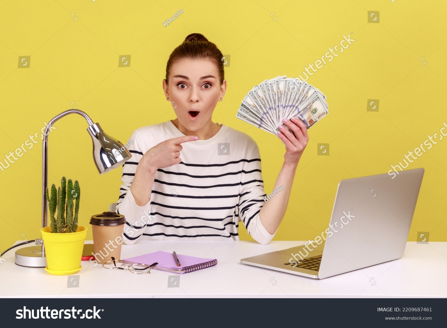 Amazed shocked woman pointing finger at hundred dollar bills, holding sitting at workplace with laptop, high salary, bonuses and perks. Indoor studio studio shot isolated on yellow background. #2209687461