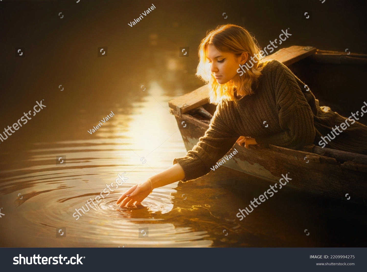  A beautiful blonde woman in a sweater sits in an old fishing boat and touches the surface of the river water with her hand, illuminated by the light of the rising sun. Tranquility and a fairy tale. #2209994275