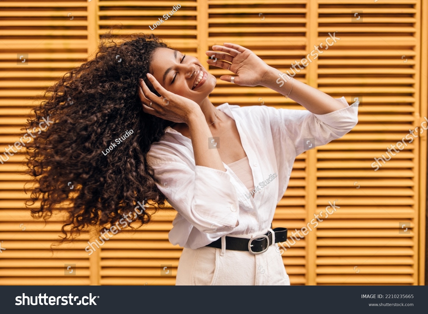 Cool young african girl waving her hair closed eyes smiling with teeth on yellow background. Model brunette curly hair wears singlet, shirt and white jeans. Concept enjoyable mood. #2210235665