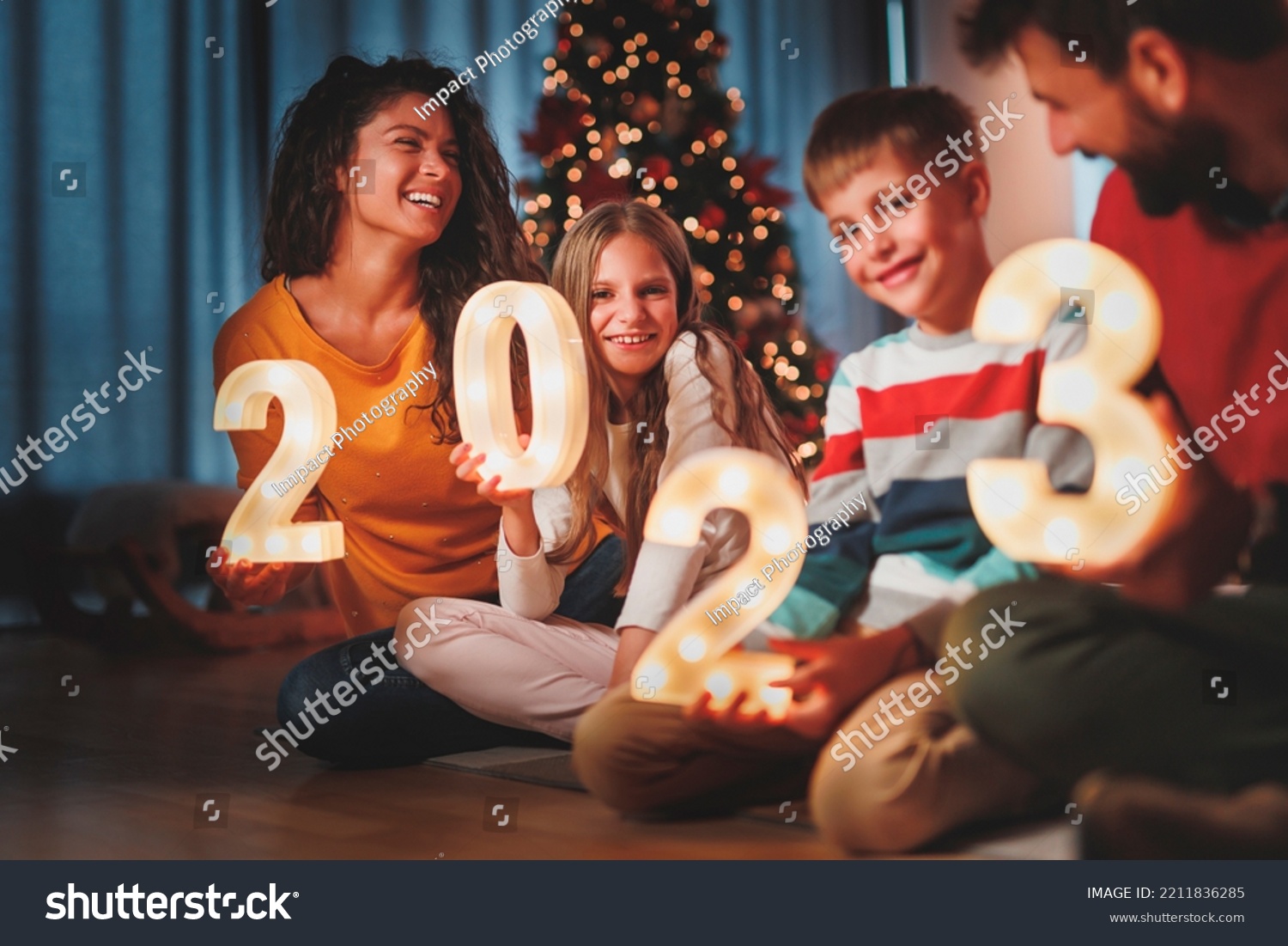 Parents celebrating New Years Eve at home with kids, sitting by the Christmas tree, holding illuminative numbers 2023 representing the upcoming New Year #2211836285