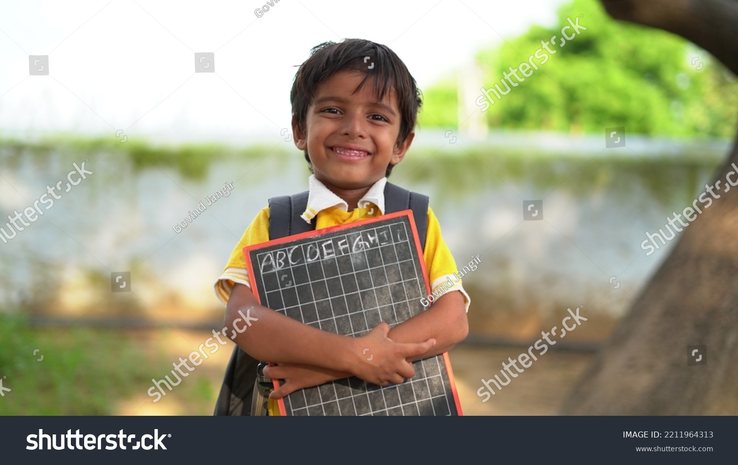 Portrait of happy cute little indian boy in school uniform holding blank slate against orange background, Adorable elementary kid showing black board. child education concept. rural india. #2211964313