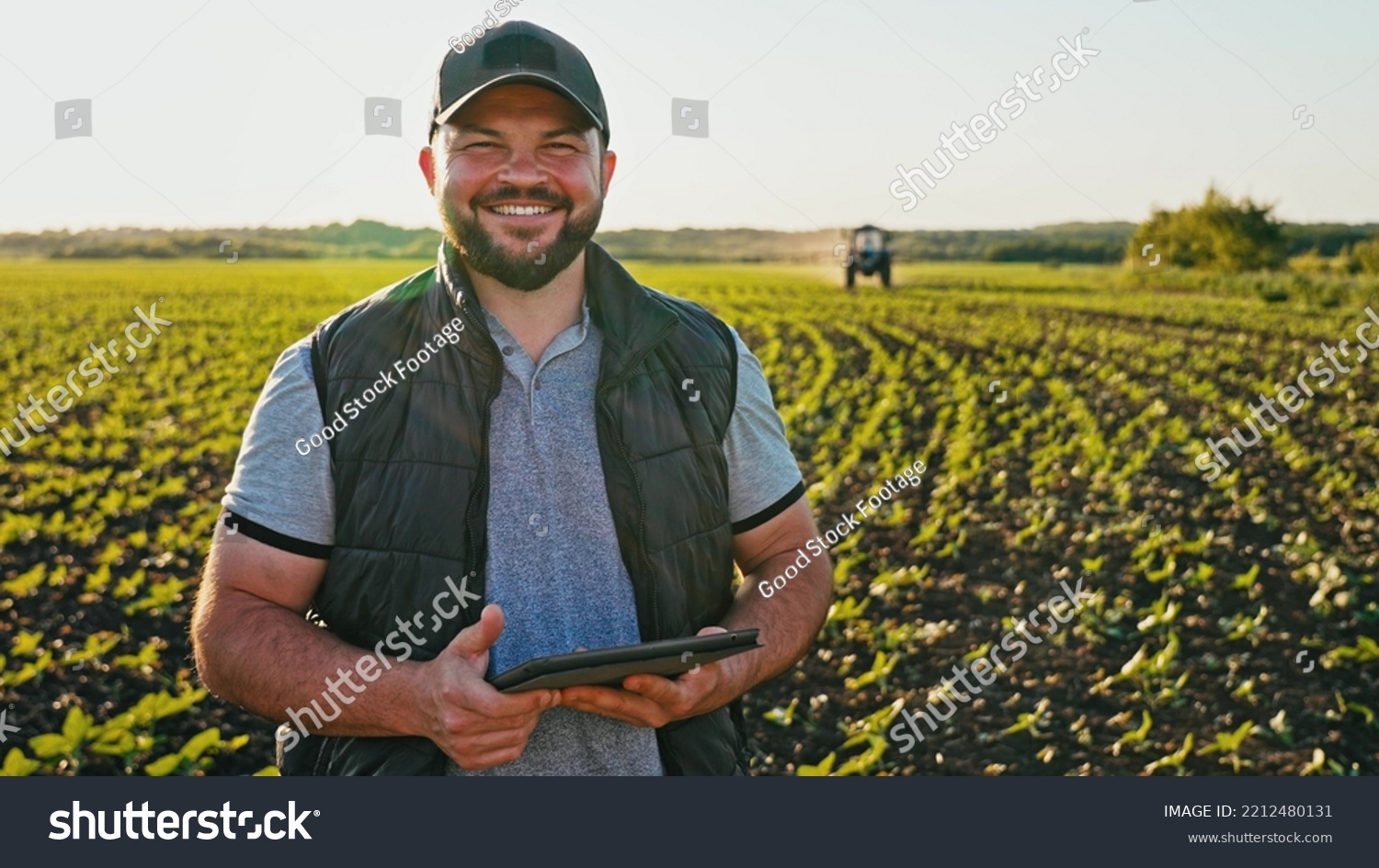 Happy farmer stands and smile holds tablet in his hands against background of working tractor in field. Concept ecology, transport, outdoor nature, clean air, food. Natural production bio product. #2212480131