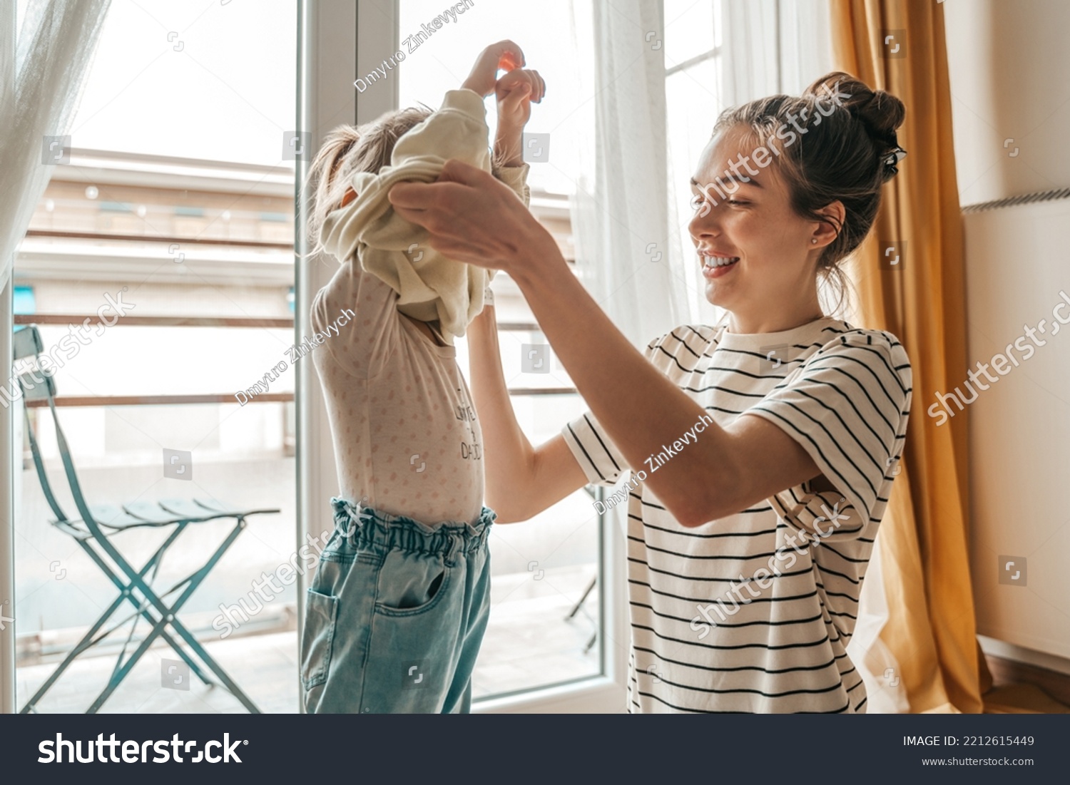Loving mother dressing her daughter by the window #2212615449