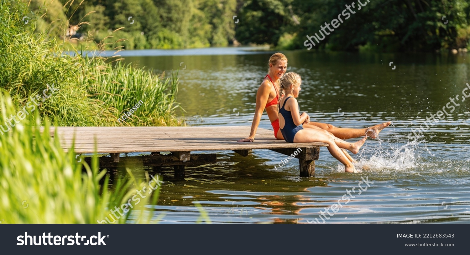 mother and daughter bathing and splashing water with their foot at jetty on a lake in summer #2212683543