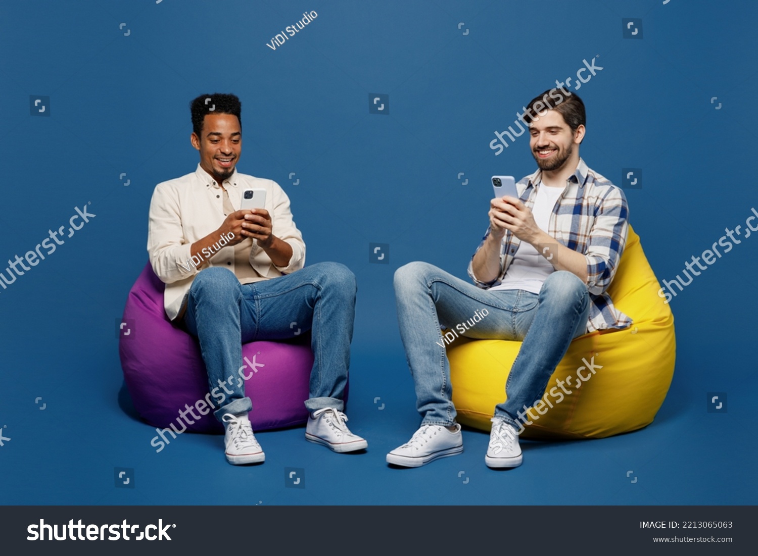 Full body young two friends happy men 20s wear white casual shirts together sit in bag chair hold in hand use mobile cell phone isolated plain dark royal navy blue background. People lifestyle concept #2213065063