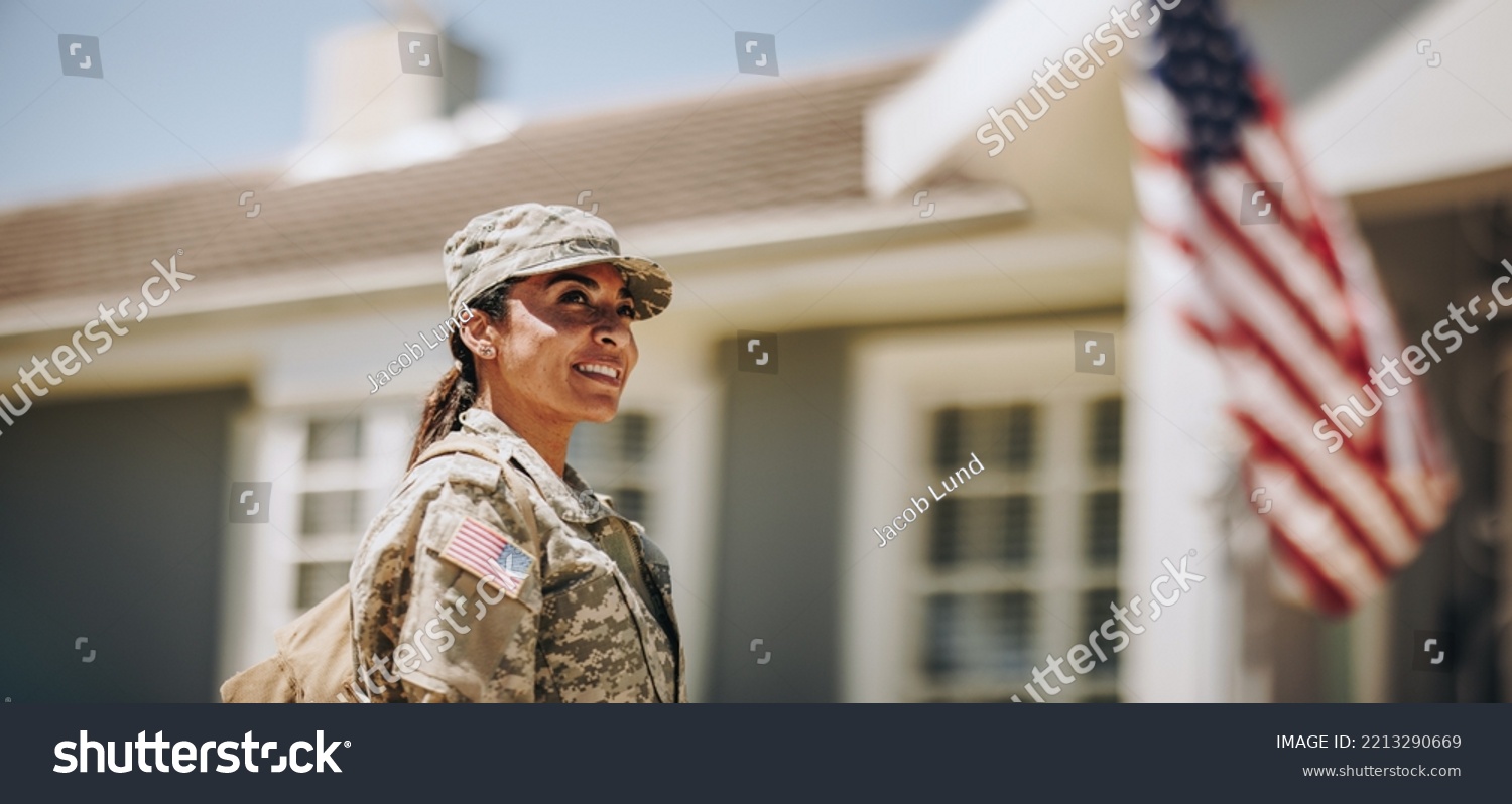 Happy female soldier looking away with a smile while standing outside her house with her bag. American servicewoman coming back home after serving her country in the military. #2213290669