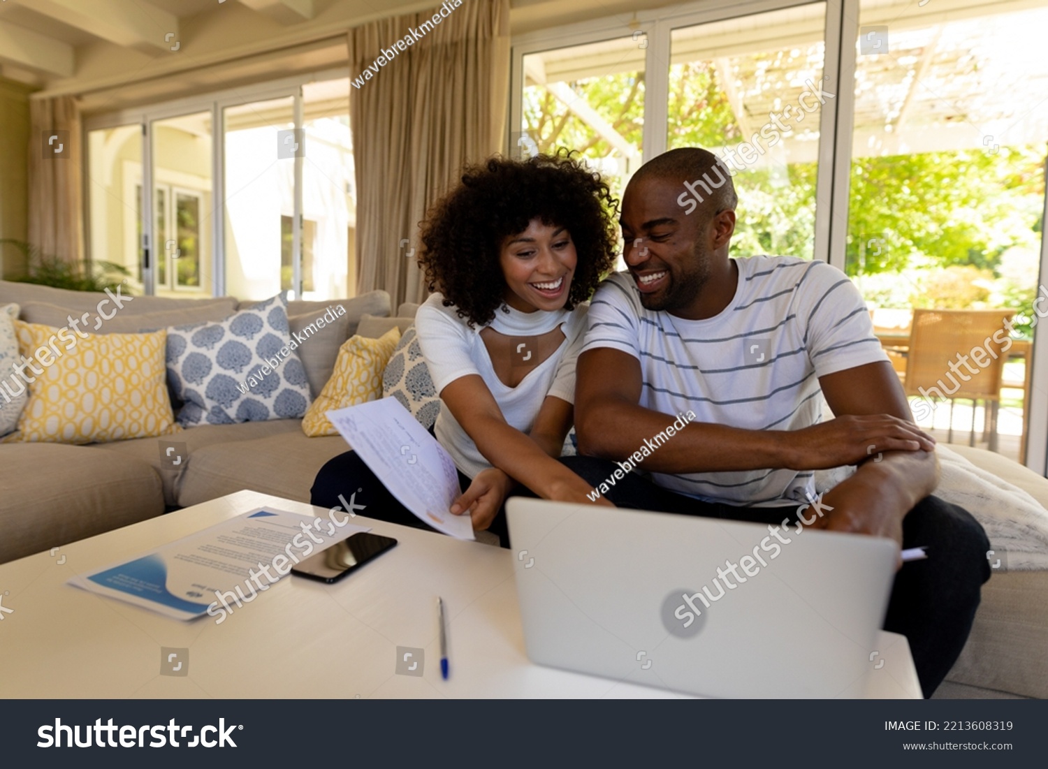 Weekend fun at home together. Front view of a mixed race couple sitting in their living room, using a laptop computer together and smiling  #2213608319
