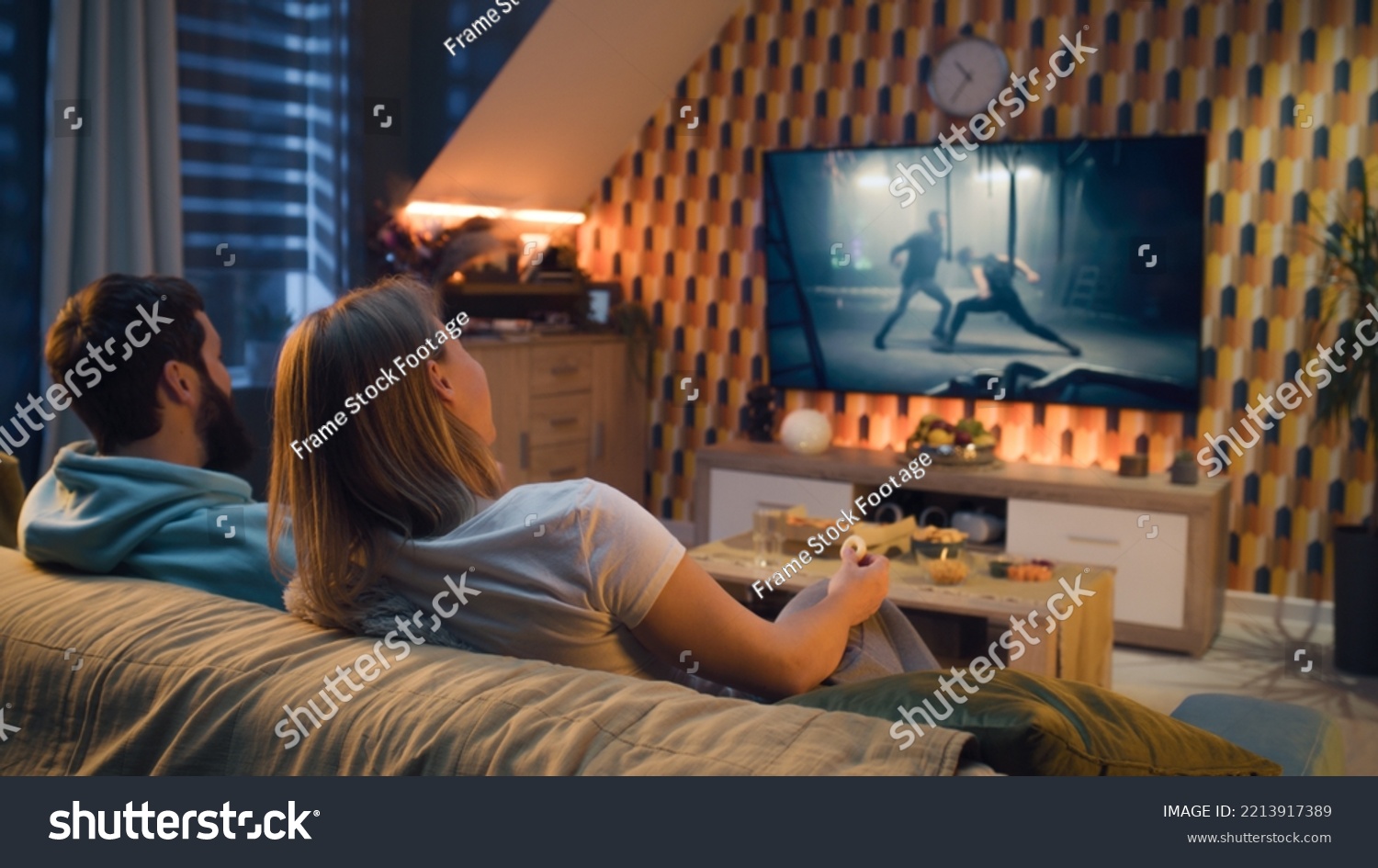 Happy couple sitting on sofa, watching action movie on TV or criminal blockbuster on streaming service, talking and discussing acting, eating snacks, pizza, chips. Spouses resting at home in evening. #2213917389